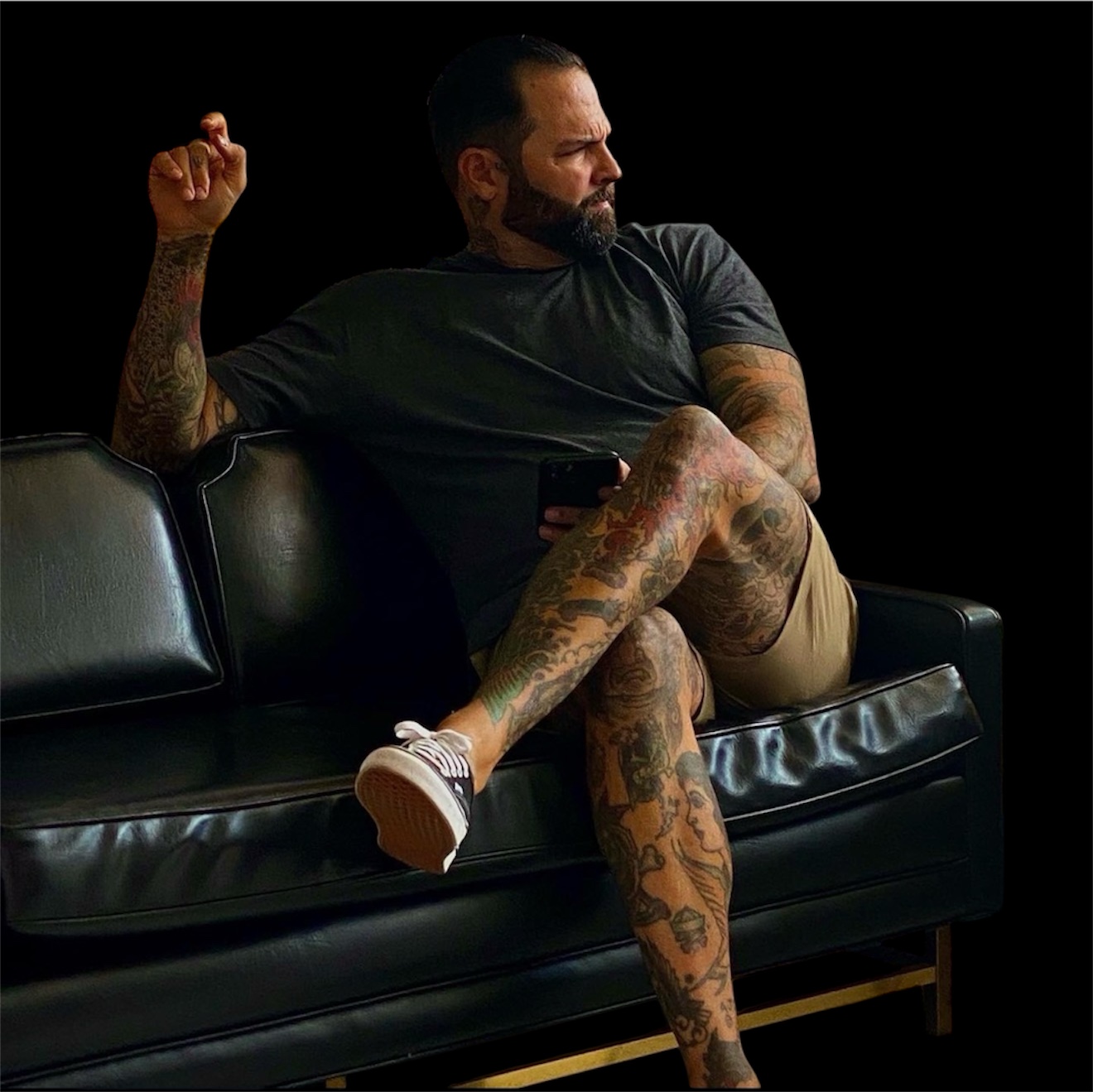 Man with tattoos sitting on a black leather couch, looking to the side, holding a phone, dark background.
