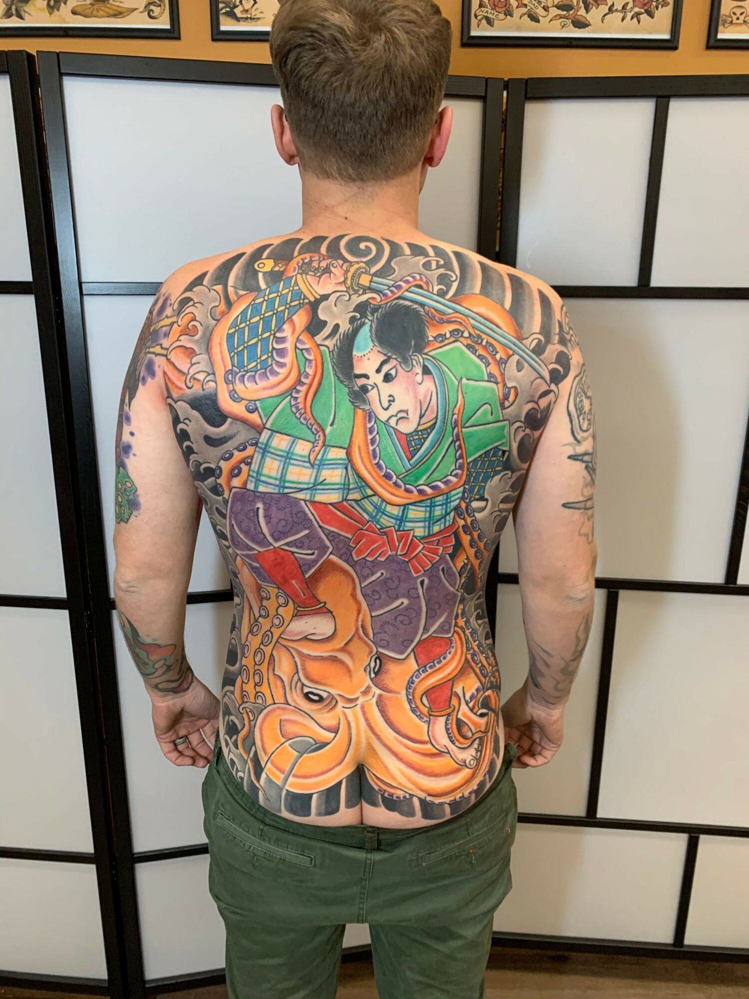 Back view of a tattooed person with colorful tattoos of a woman, dragon, and tiger on their back, standing in front of a black and white screen.