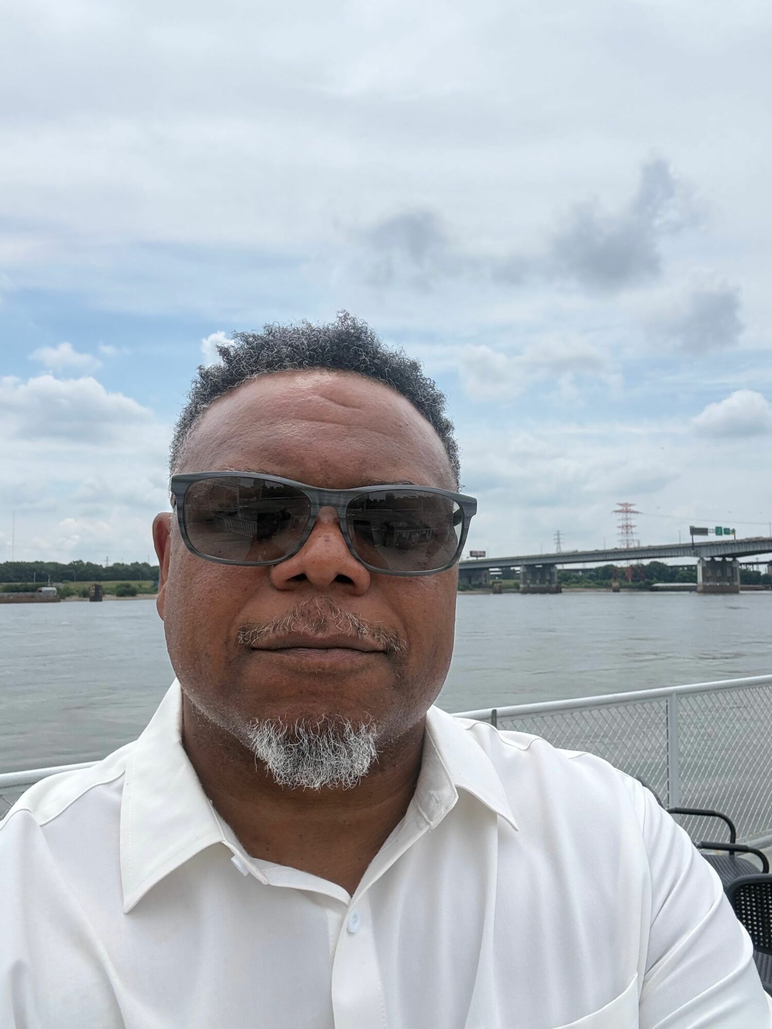 Man wearing sunglasses and white shirt outdoors near water, cloudy sky in background.