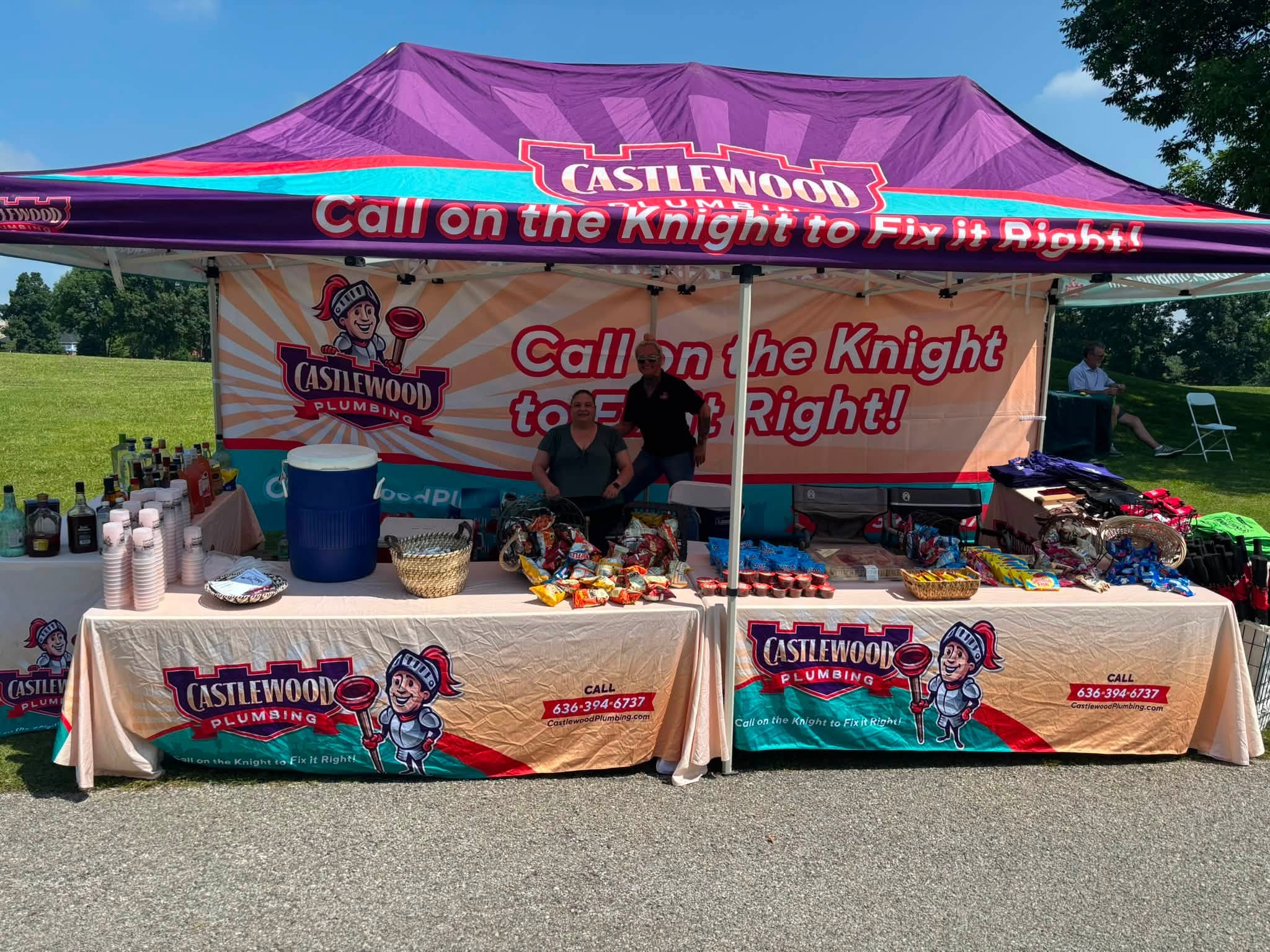 Two people sit behind a table under a purple tent with Castlewood branding, selling snacks and drinks outdoors.