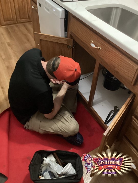 Person kneeling on red carpet, working under kitchen sink cabinet, with open cabinet door and toolbox nearby.