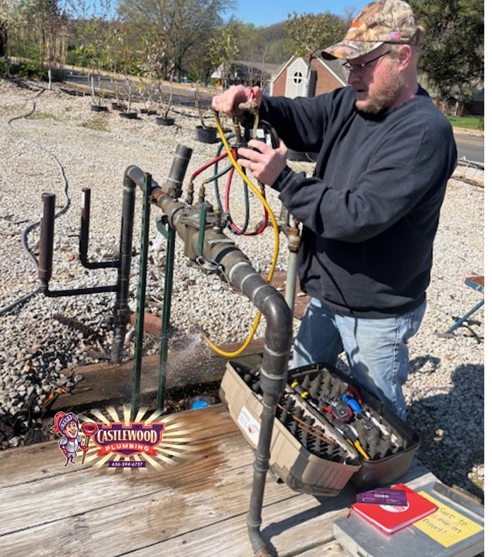 Man in camouflage hat and dark clothing working with pipes outdoors, toolbox nearby, water flowing from pipes, gravel ground, trees in background.