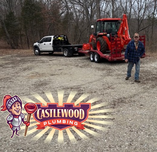Person standing outdoors near a red tractor on a trailer, with a pickup truck in the background, trees, and a Castlewood Plumbing sign.
