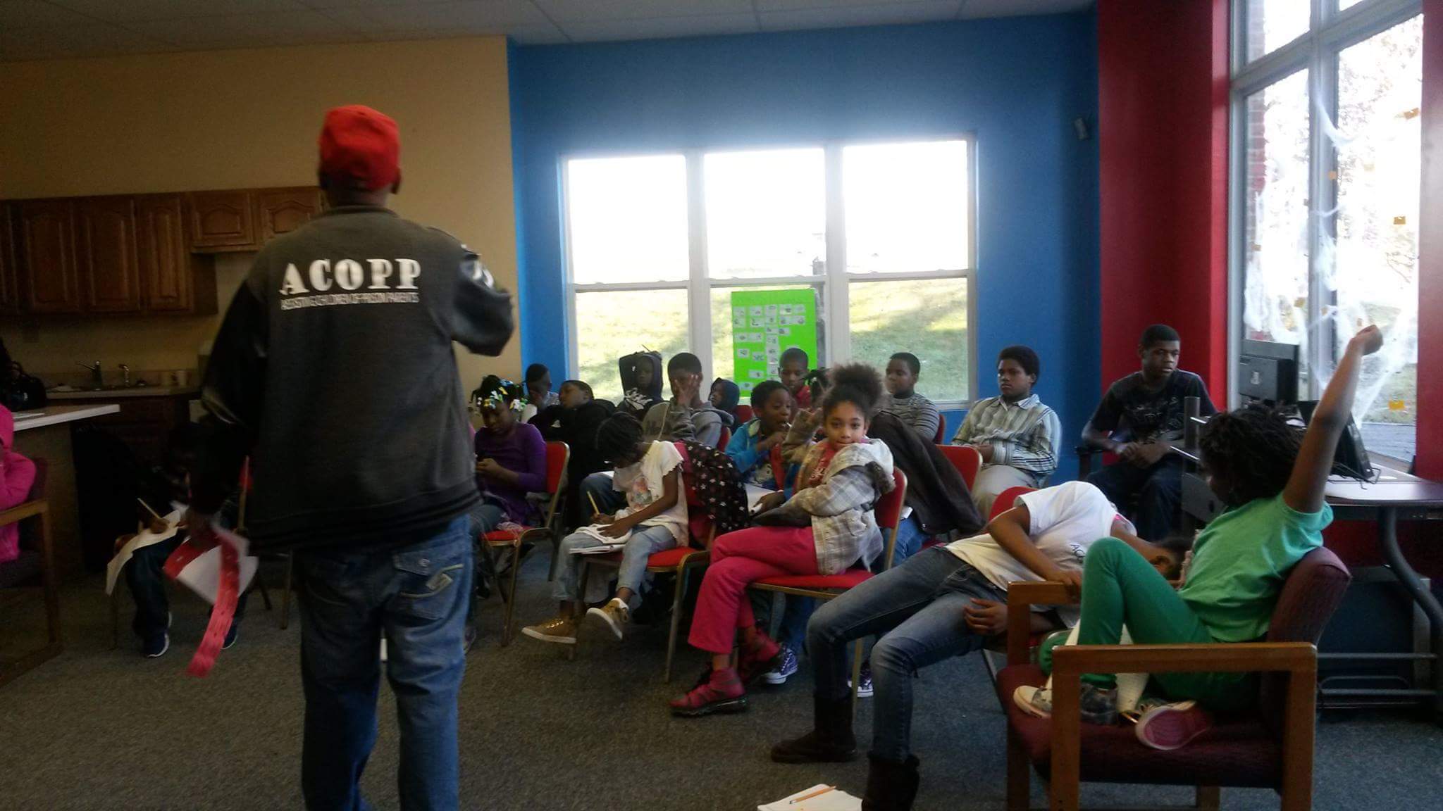 A group of children seated in a classroom with large windows, some raising hands, and a person standing at the front.