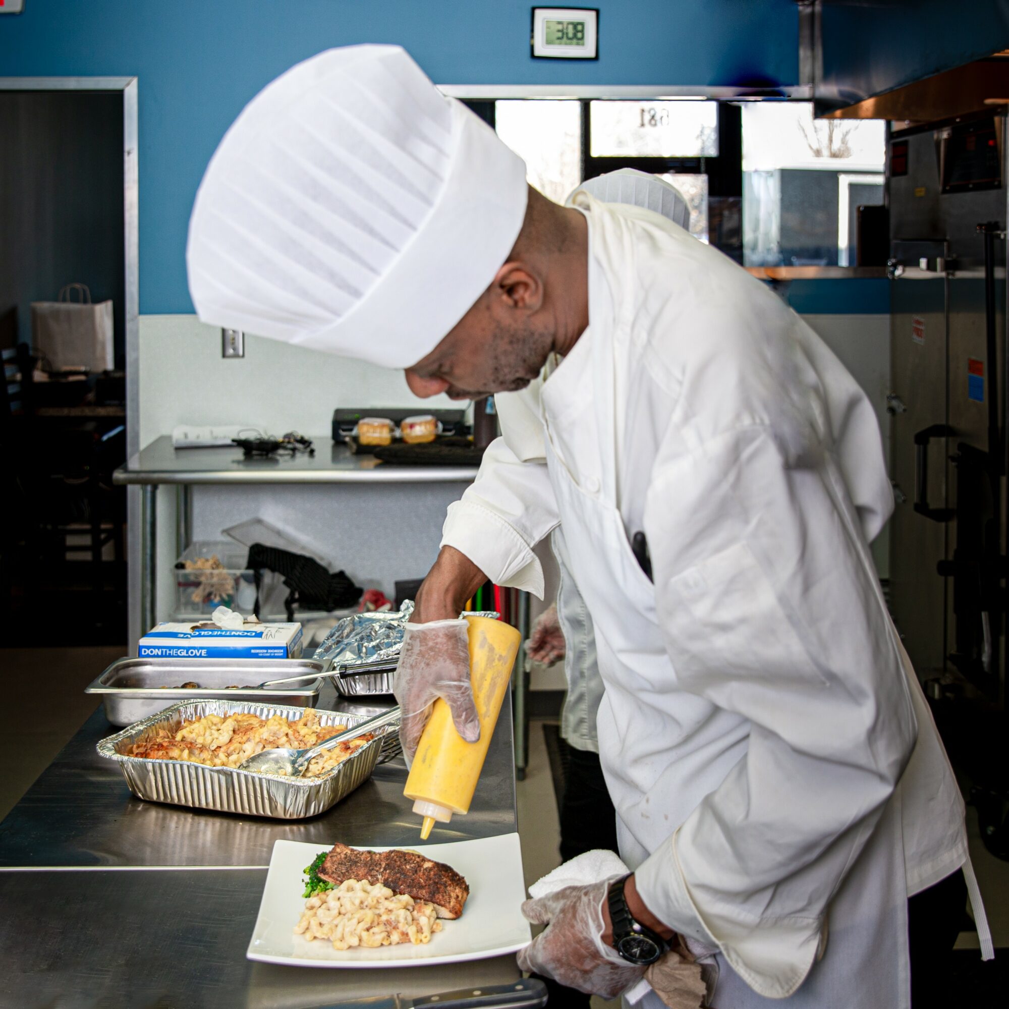 Chef in white uniform and hat preparing food with mustard on a plate in a kitchen.