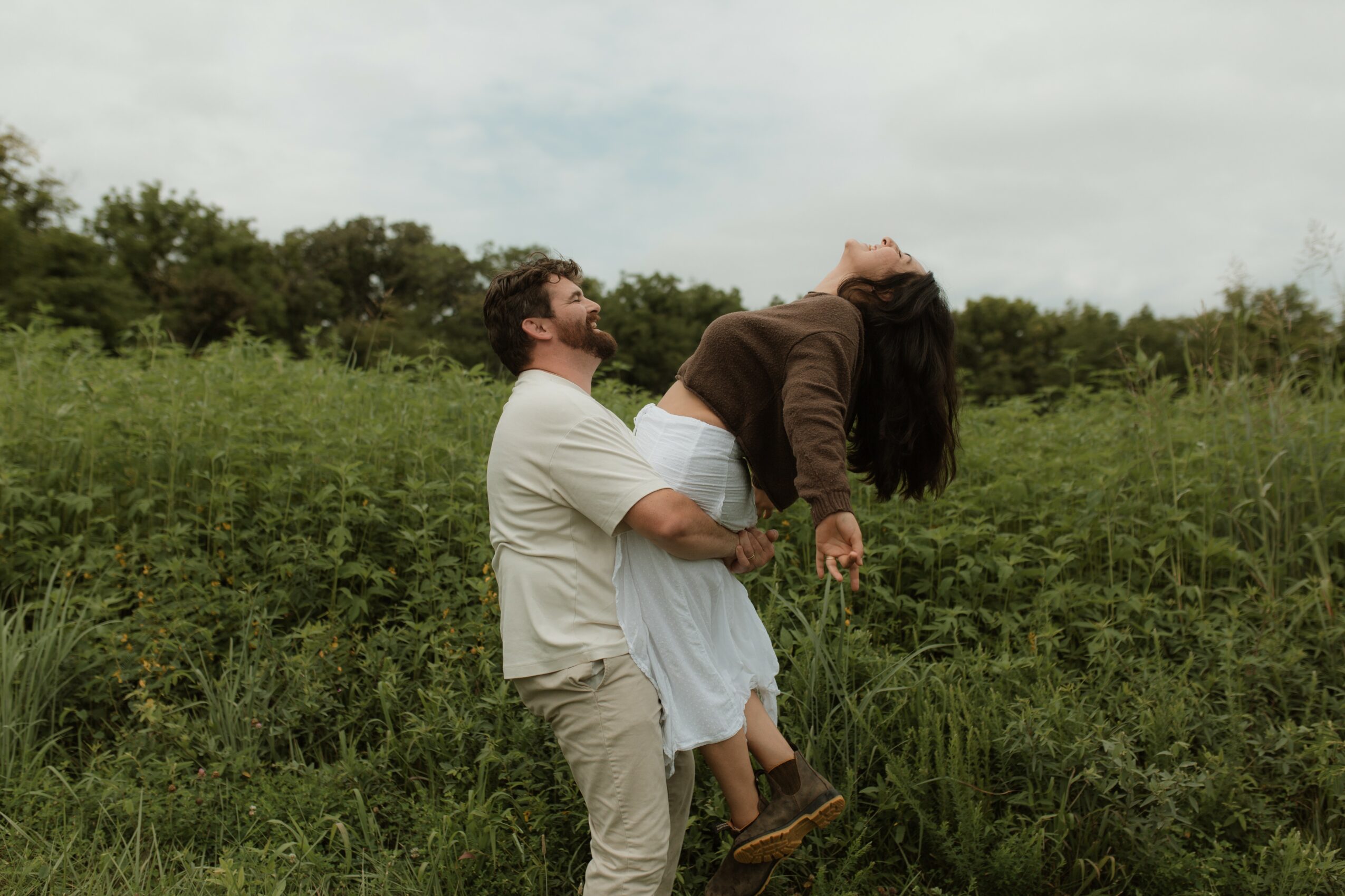 Man holding woman in a field with greenery and trees in the background, both smiling and relaxed.