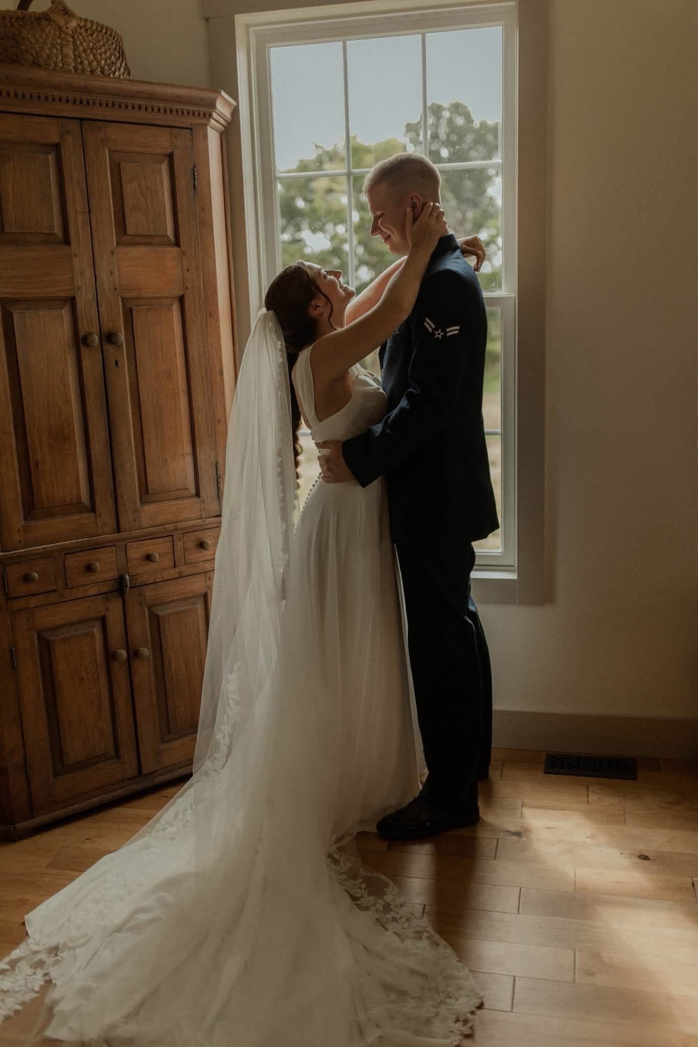 Bride and groom embrace indoors near a window, with wooden furniture in the background.