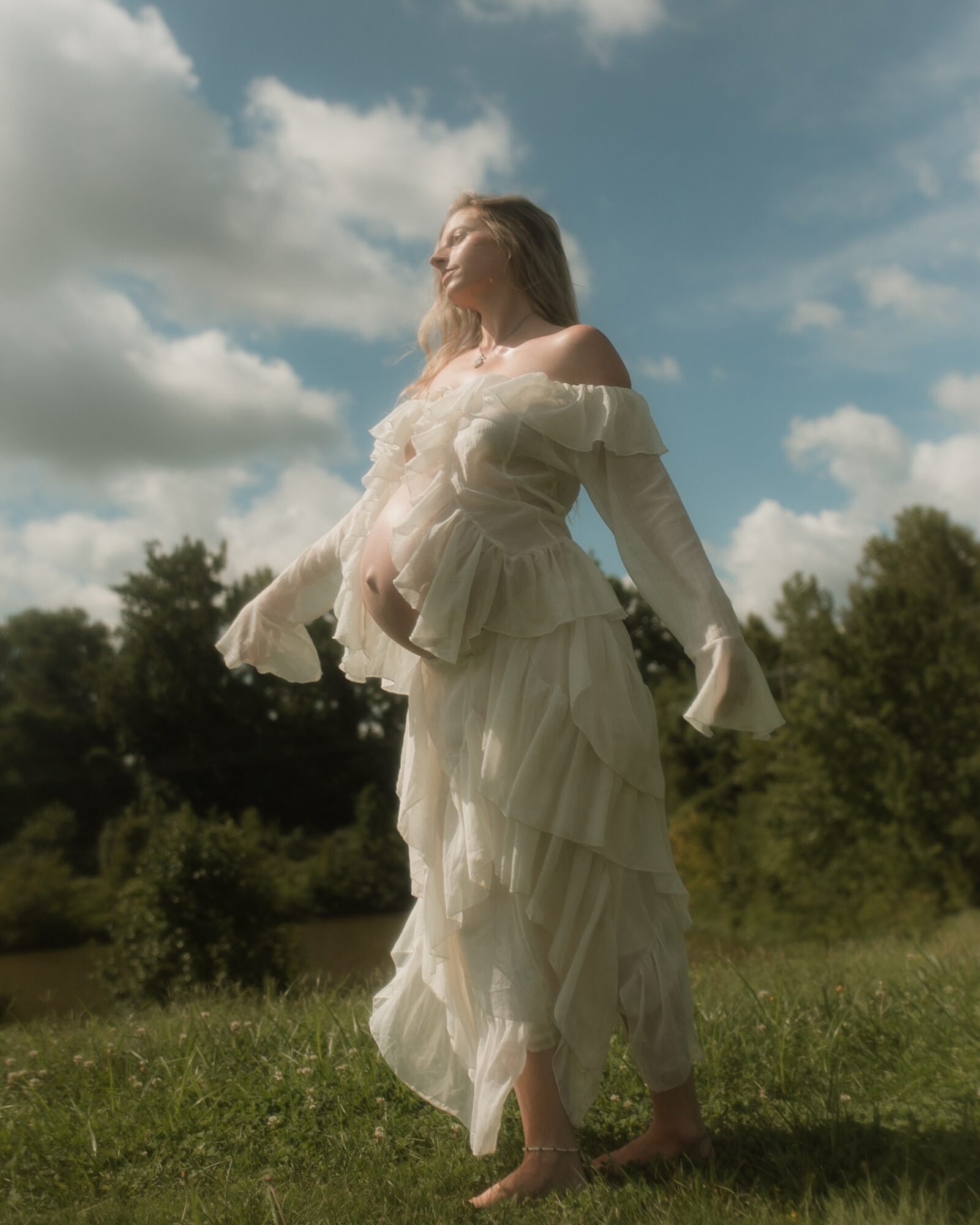 Woman in a flowing white dress standing outdoors on grass, looking up at the sky with clouds, trees in background.