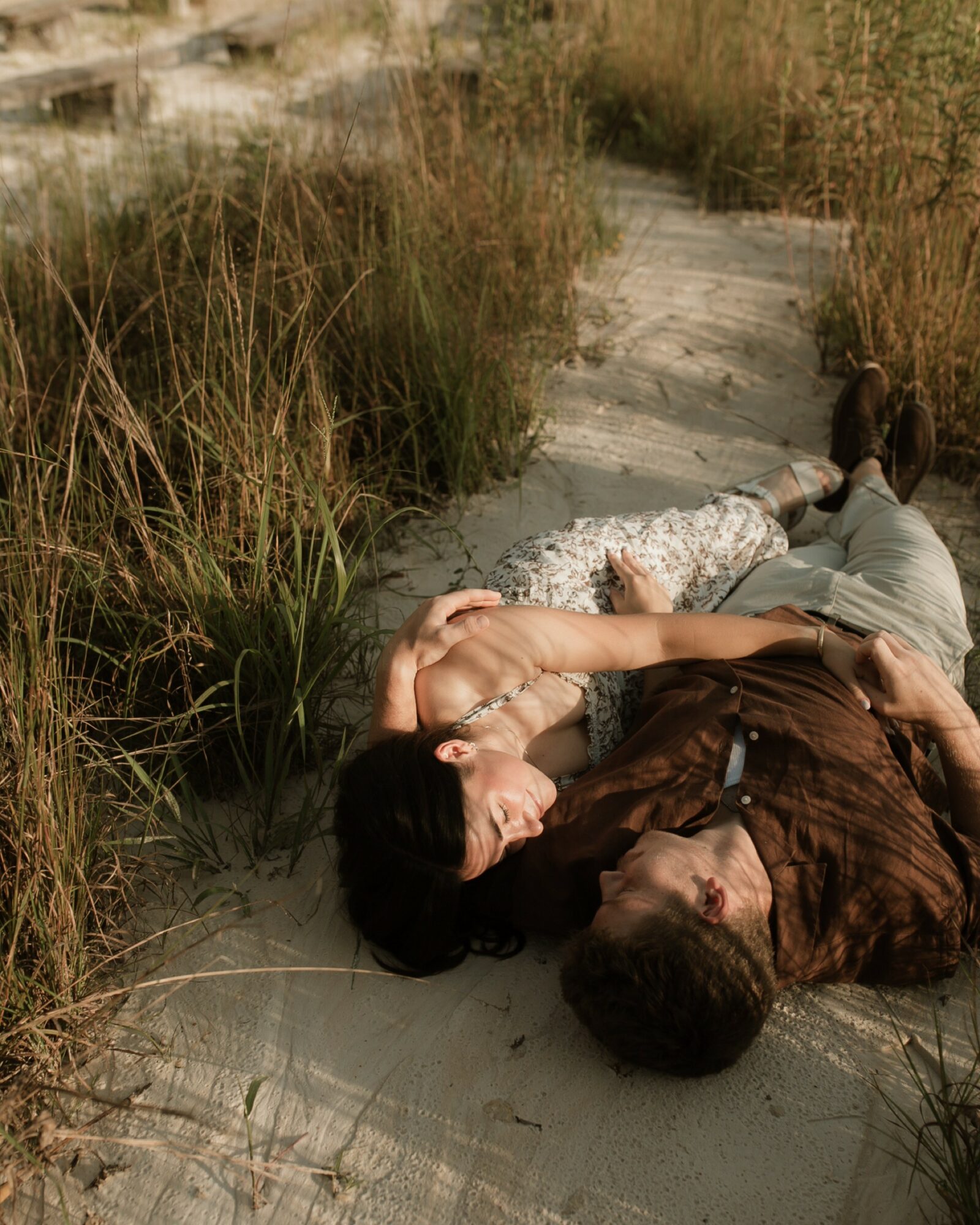 Two people lie on a sandy path surrounded by tall grass, resting outdoors on a sunny day.