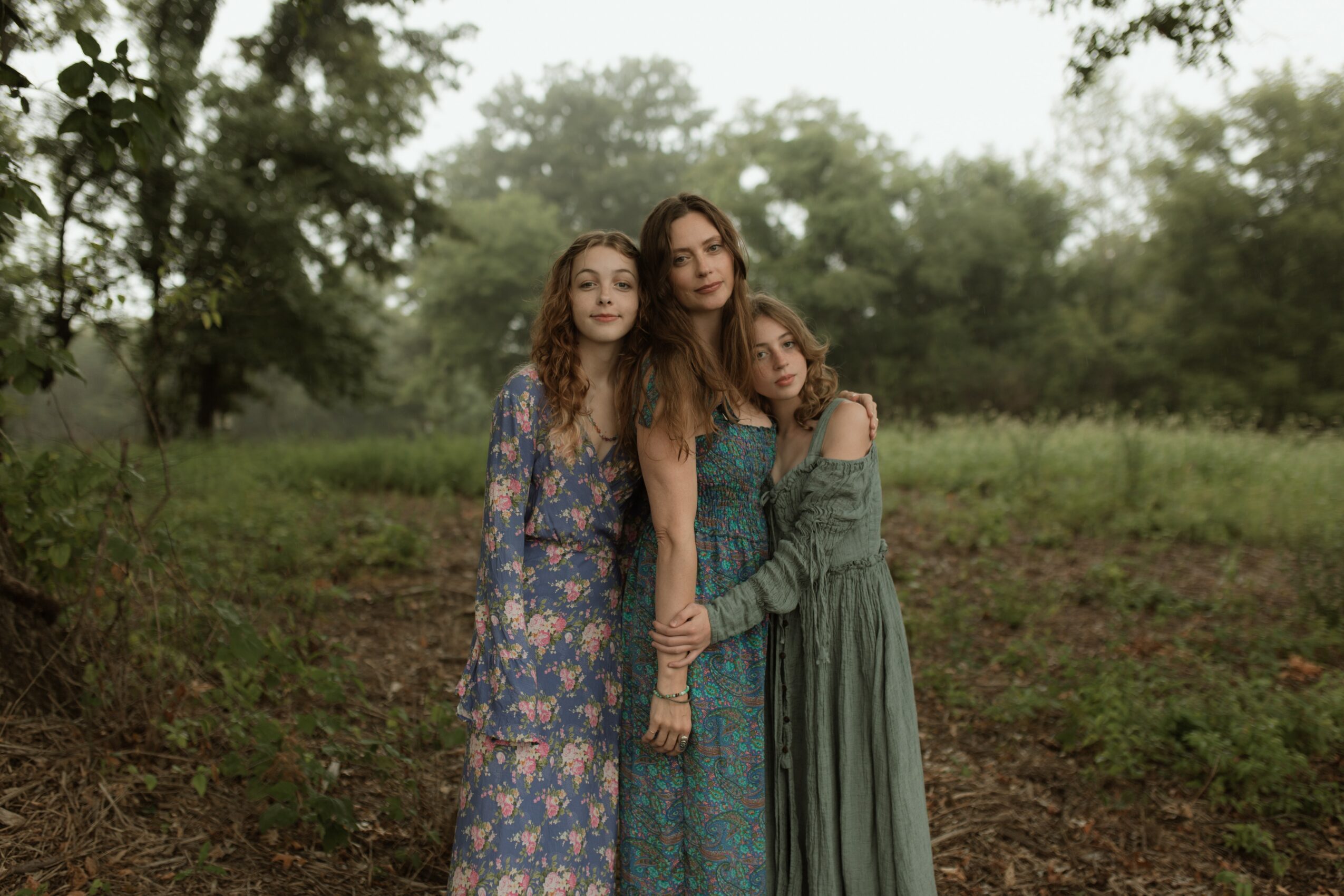 Three young women standing outdoors in a grassy area with trees, wearing patterned dresses, holding hands, and looking at the camera.