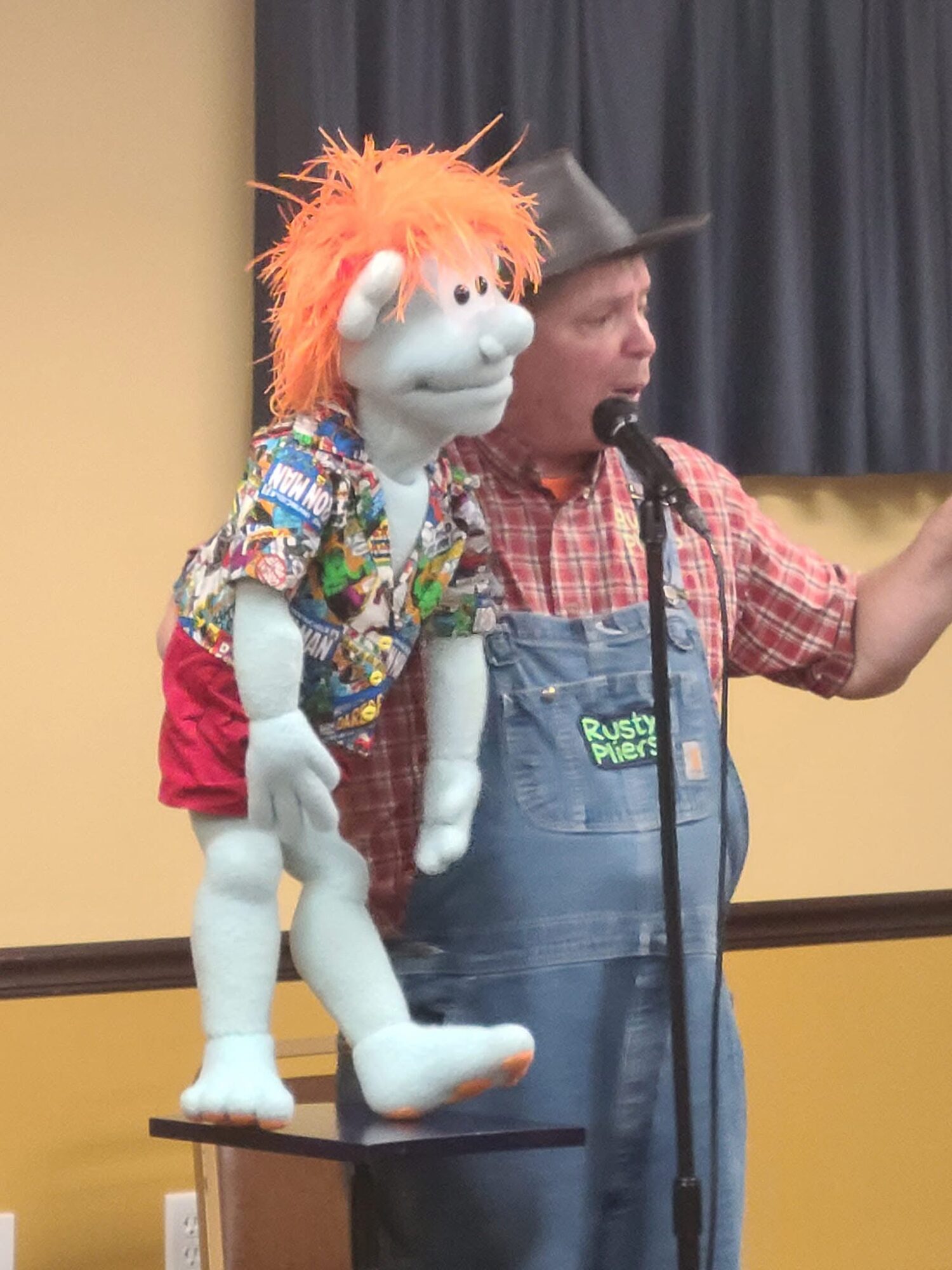 A man wearing a cowboy hat and plaid shirt holds a puppet with orange hair and a colorful shirt, standing on a table.