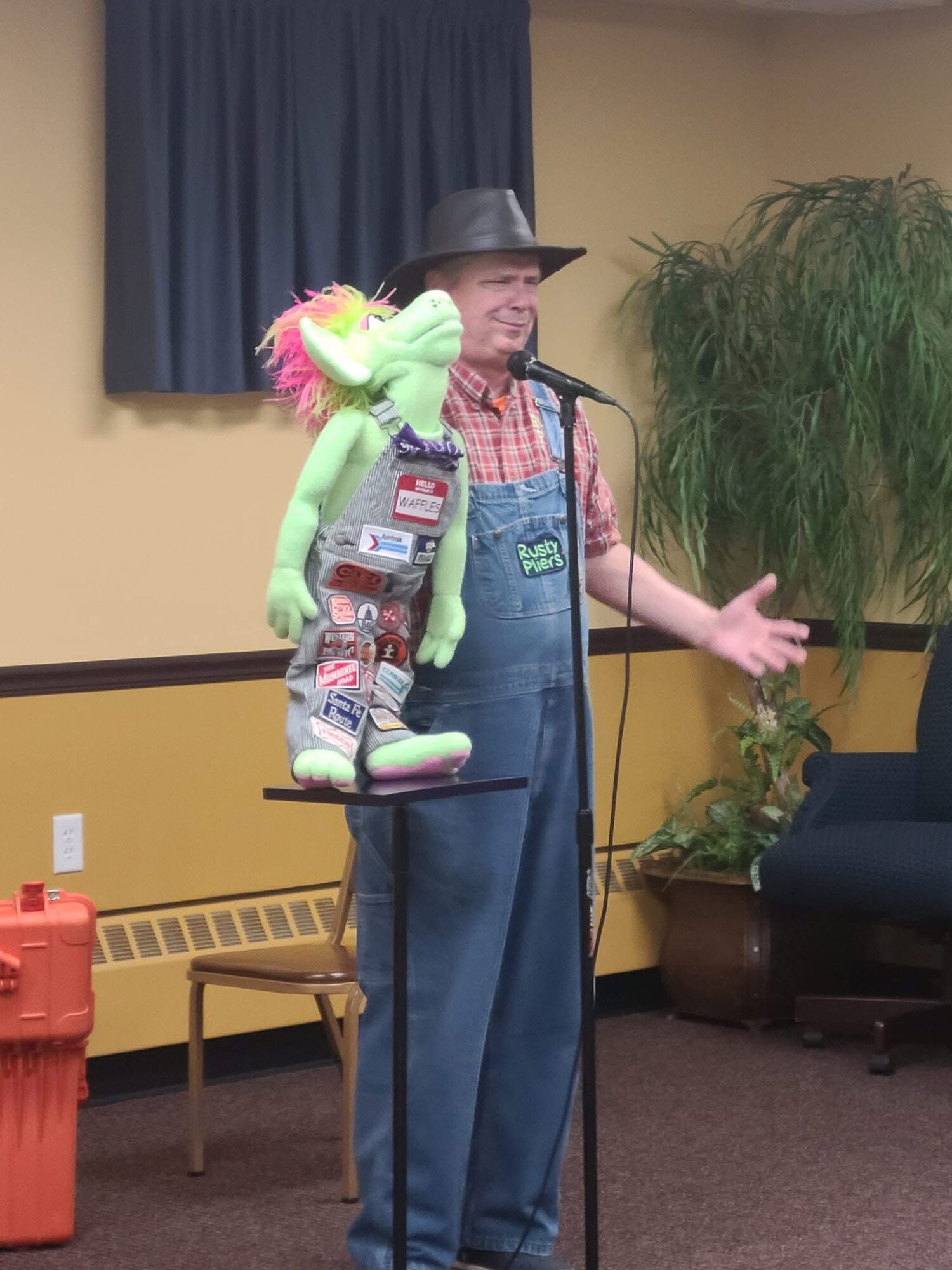 Man wearing a hat and overalls stands at microphone with puppet on his shoulder, in a room with plants and furniture.