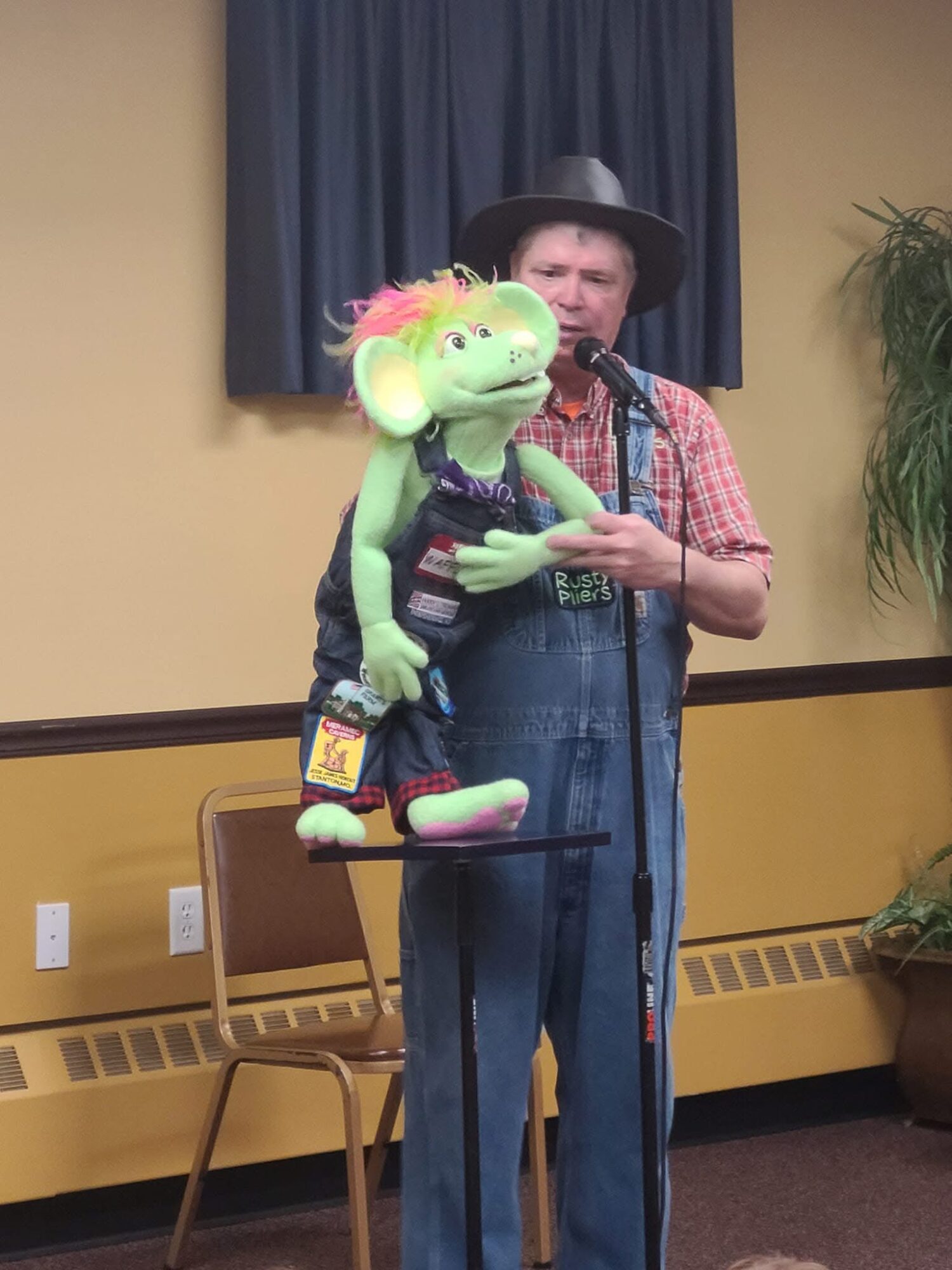 Person wearing a hat and plaid shirt holding a puppet with a microphone in a room with yellow walls and a blue curtain.