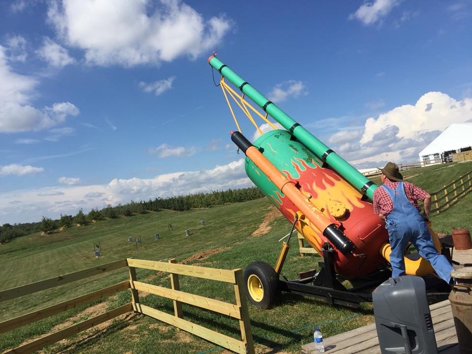 Person in overalls adjusting a large colorful cannon-like structure outdoors, with a wooden fence and grassy field in background.