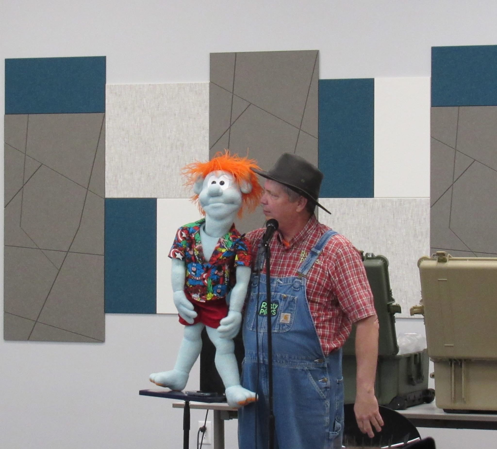 A puppet with orange hair and a colorful shirt stands on a table next to a man in overalls and a hat, speaking into a microphone.