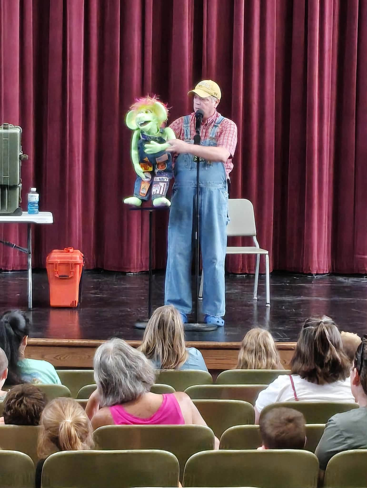 Performer with a puppet on stage in front of red curtains, audience watching, chair and trash bin nearby.