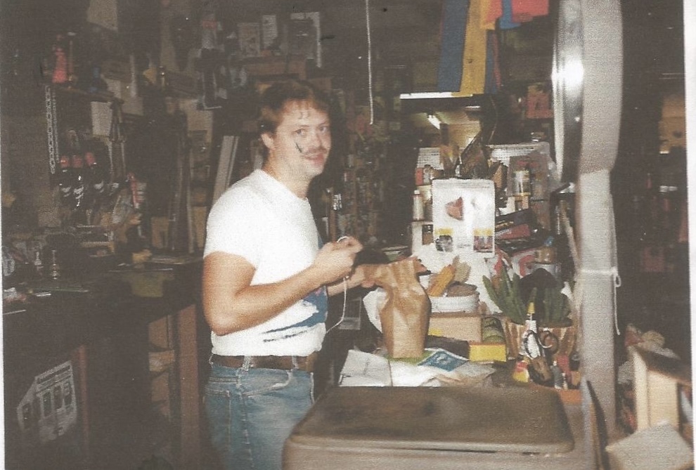 Person smiling and holding a bag in a cluttered store, surrounded by shelves and items.