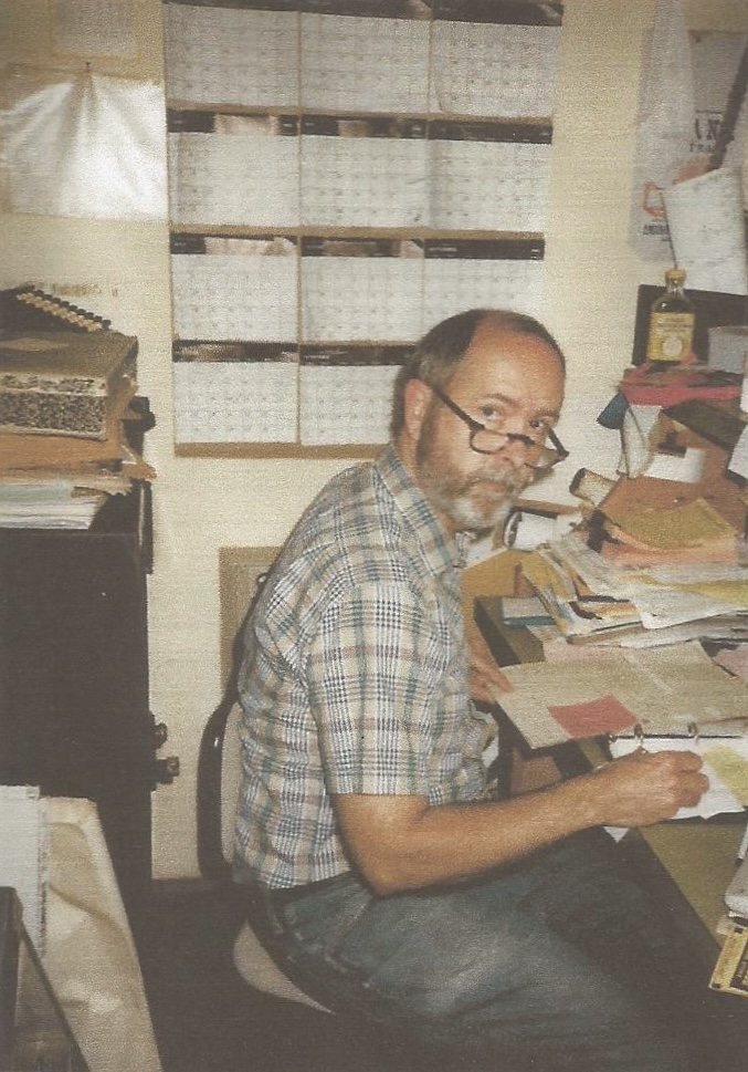 Man with glasses and a checkered shirt sitting at a cluttered desk, smiling, in an office with shelves and papers.