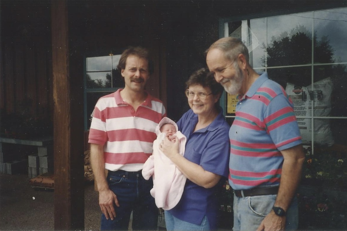 Three people standing outside a building, one holding a newborn baby, smiling and looking at the baby.