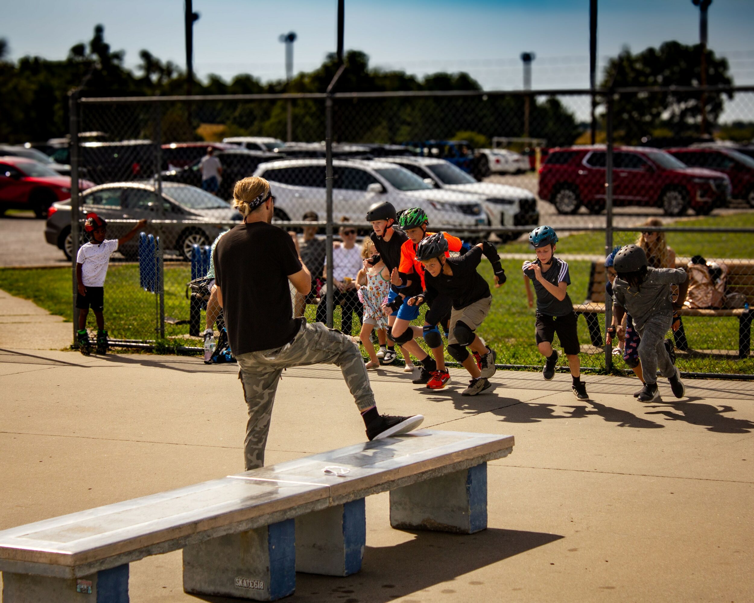 Children skateboarding and playing near a fence at a park, with spectators and parked cars in the background.