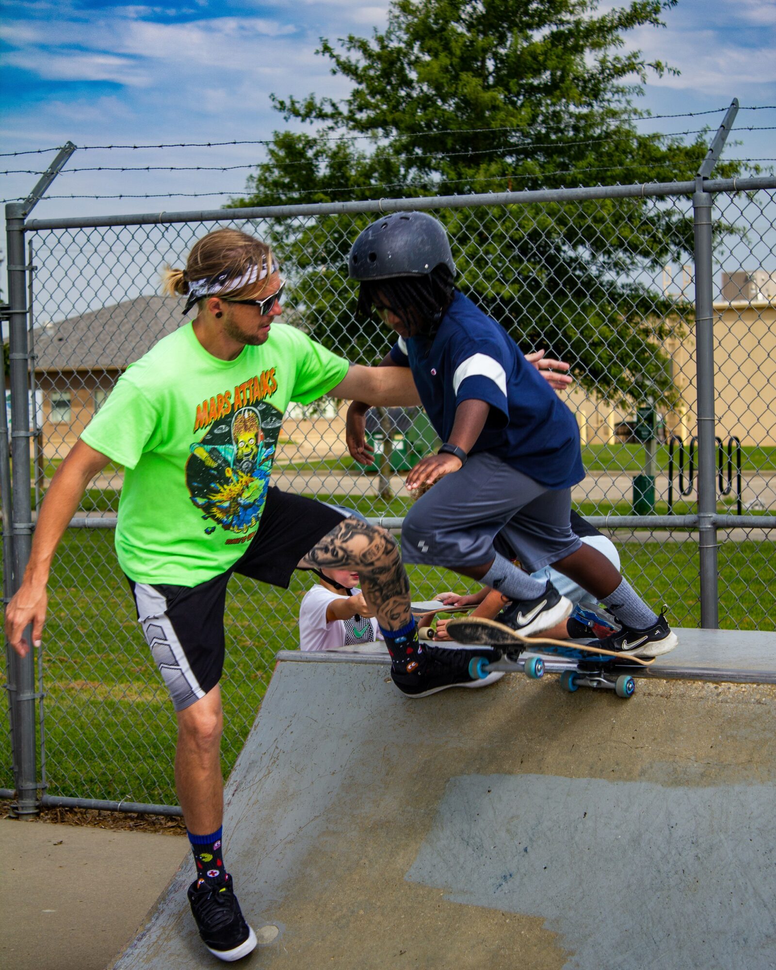 Child in a helmet skateboarding on a ramp while a person in a bright green shirt assists, outdoors with a fence and trees.