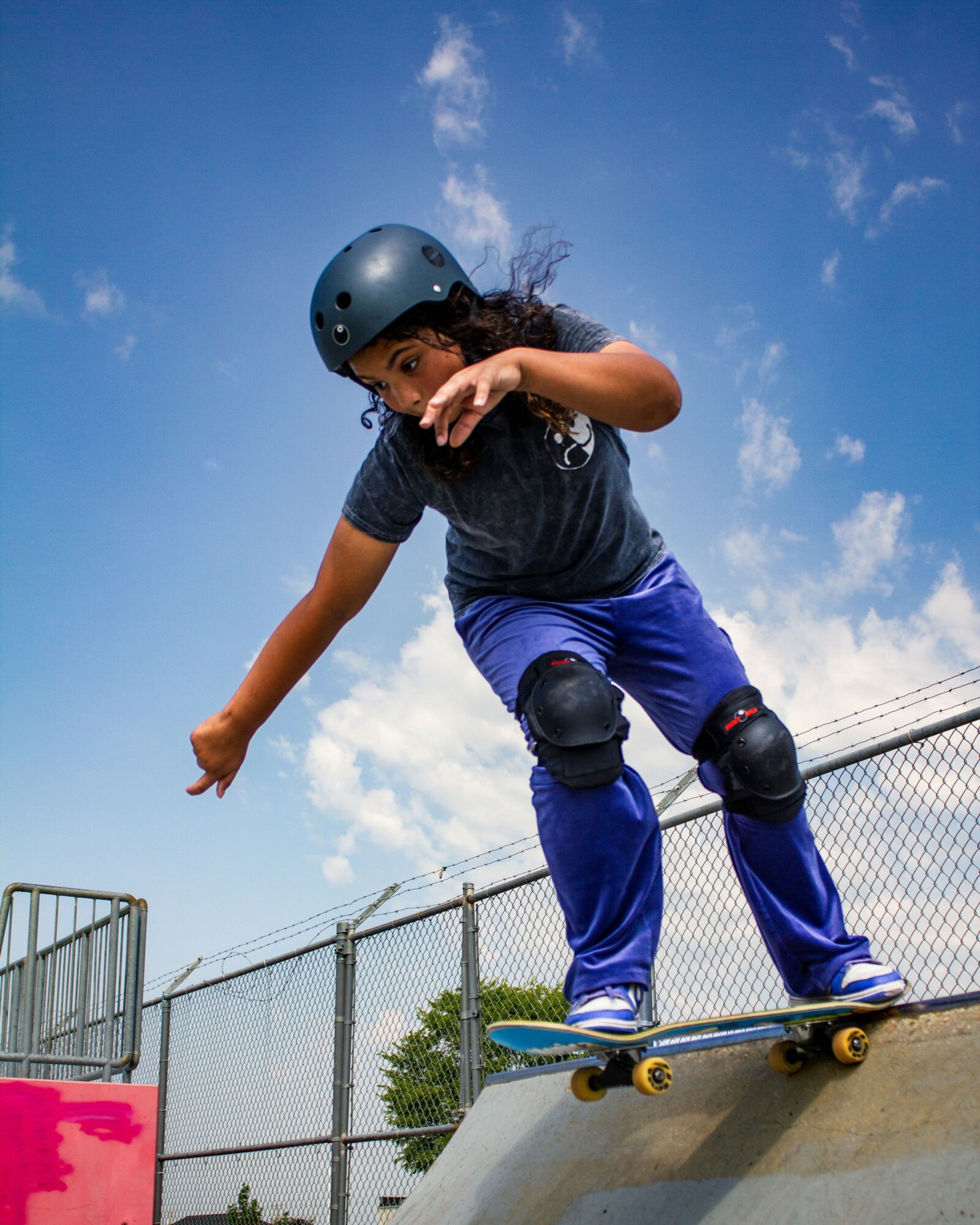 Child wearing a helmet skateboarding on a ramp outdoors, with a fence and blue sky in the background.