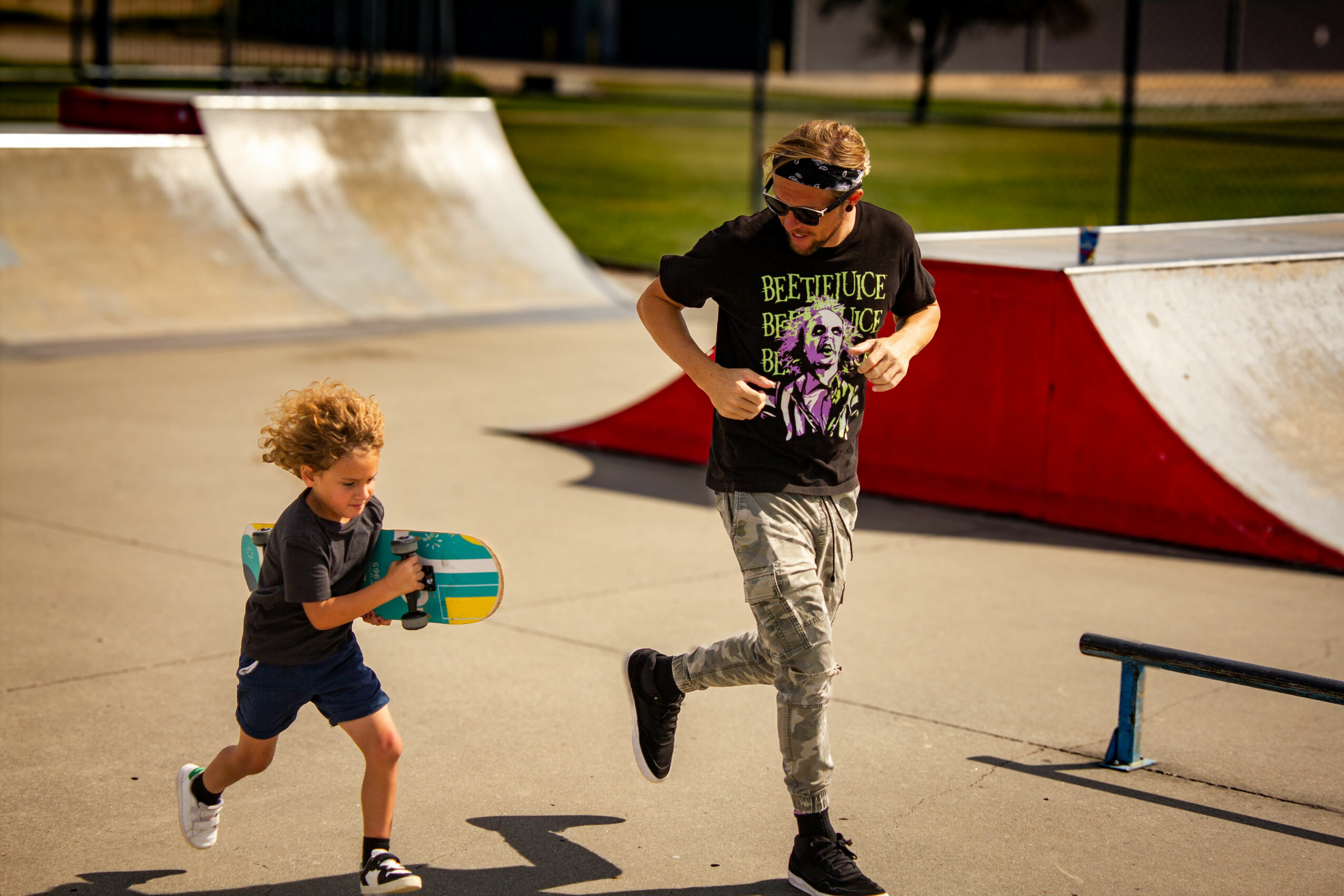 Two children running at a skate park, one holding a skateboard, with ramps in the background.