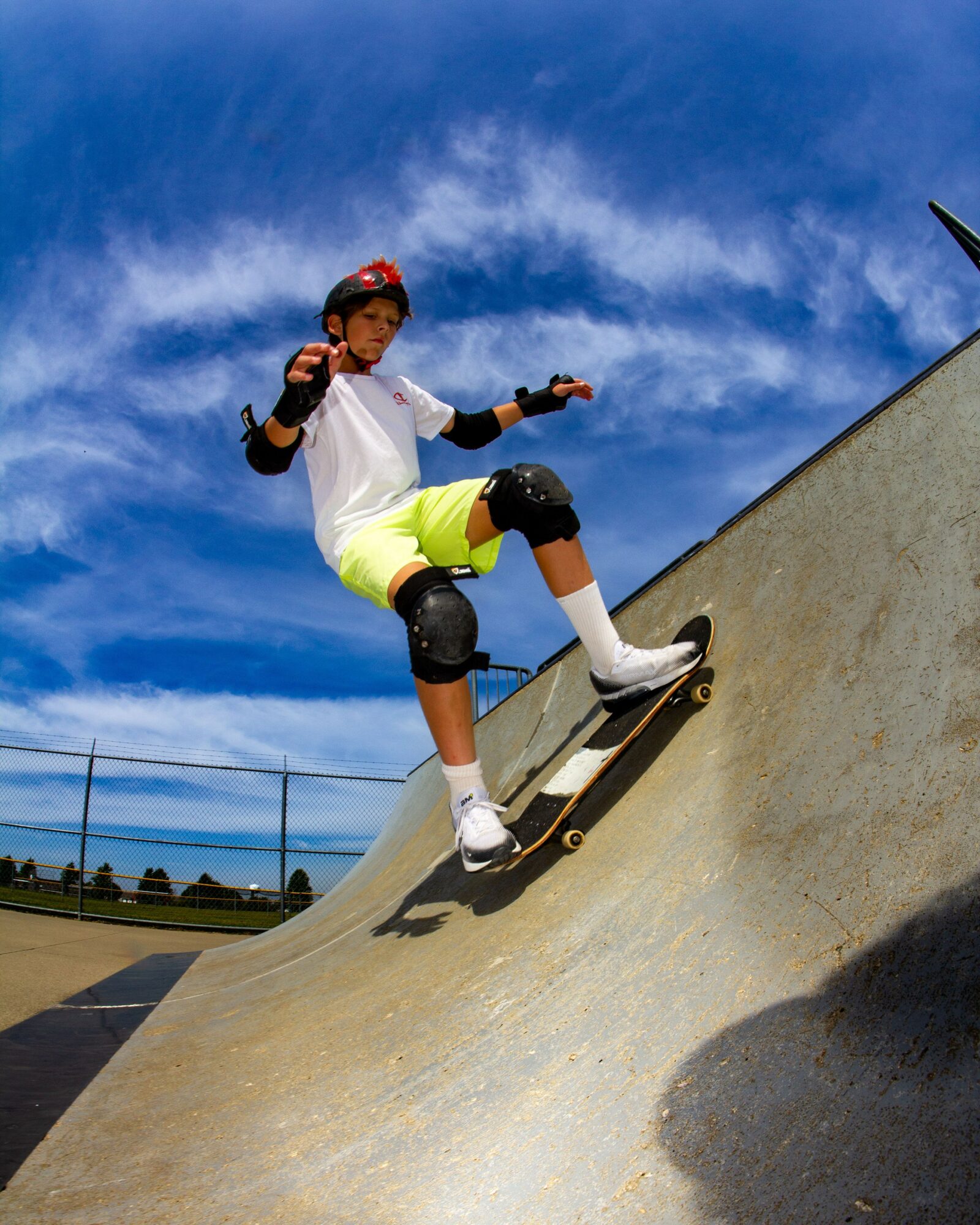 A young person skateboarding on a ramp outdoors under a blue sky with clouds, wearing a helmet and protective gear.