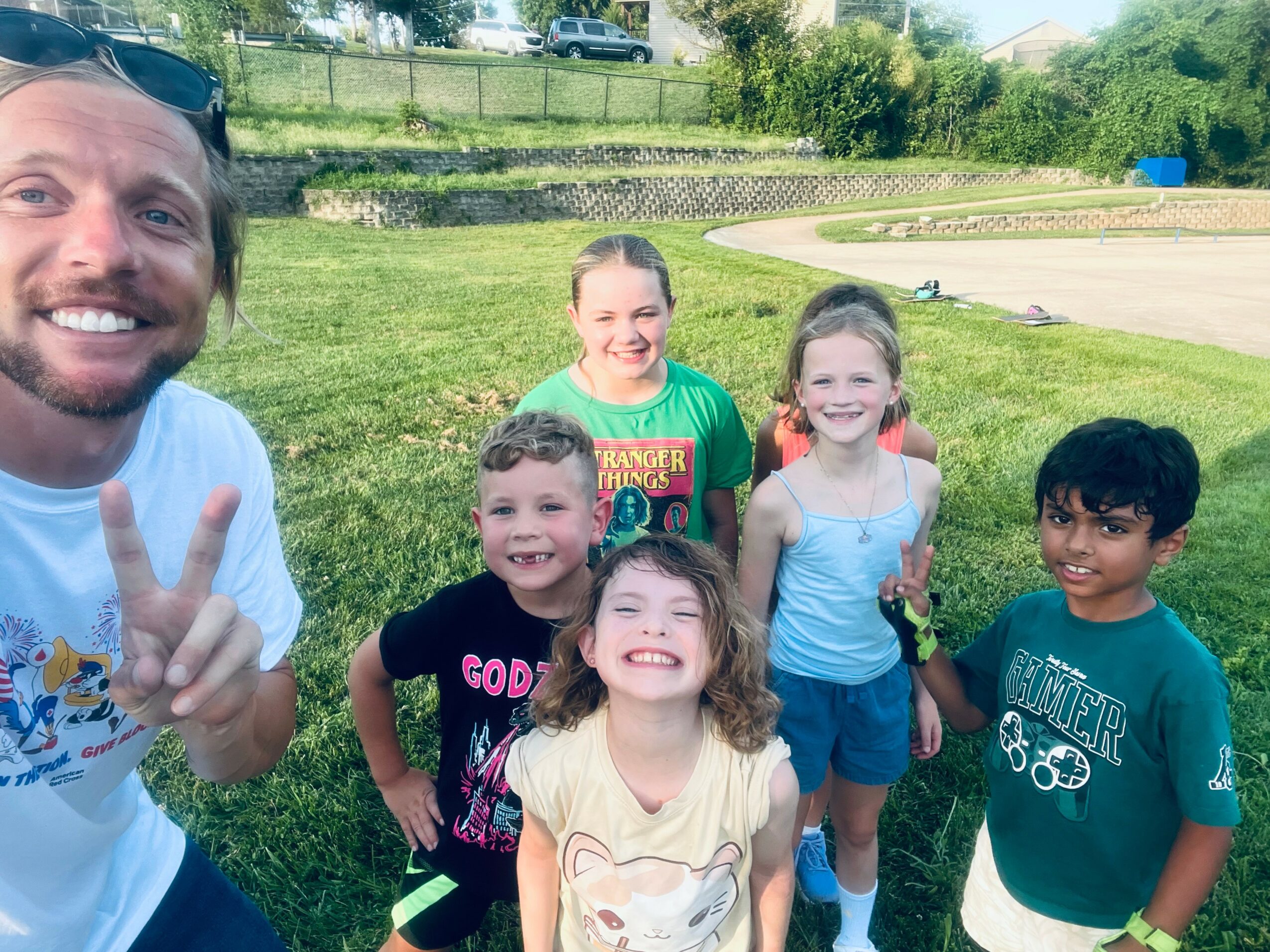 Group of five children and one adult smiling outdoors on grass, with trees and a fence in background.