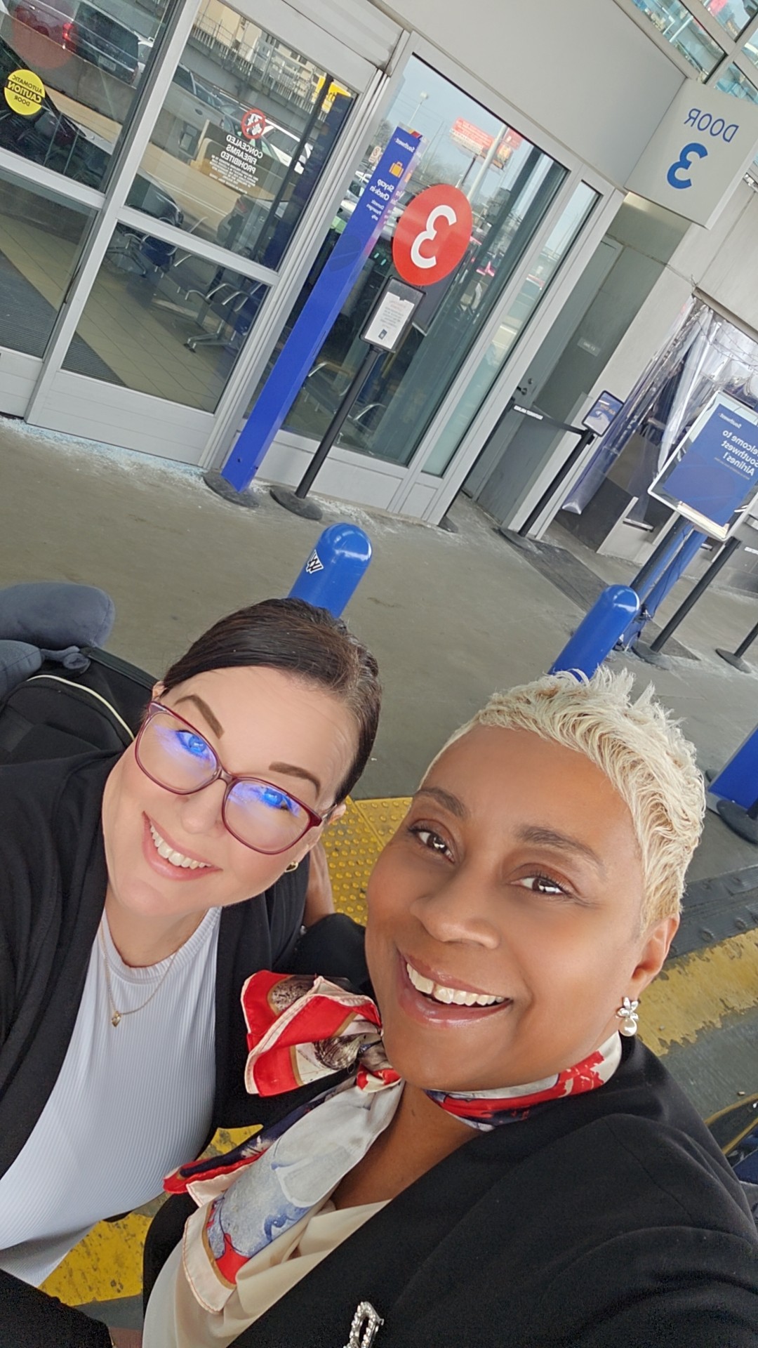 Two women smiling outside a building with glass doors, one with glasses and short dark hair, the other with short blonde hair and a scarf.