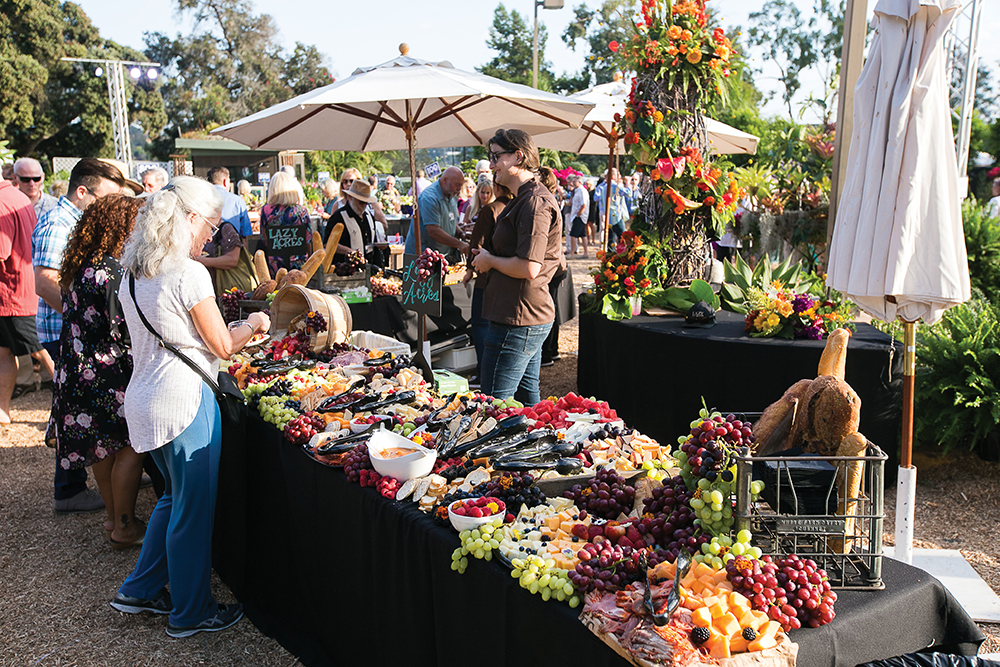 People browsing a market stall with jewelry and flowers outdoors on a sunny day, with umbrellas and trees in the background.