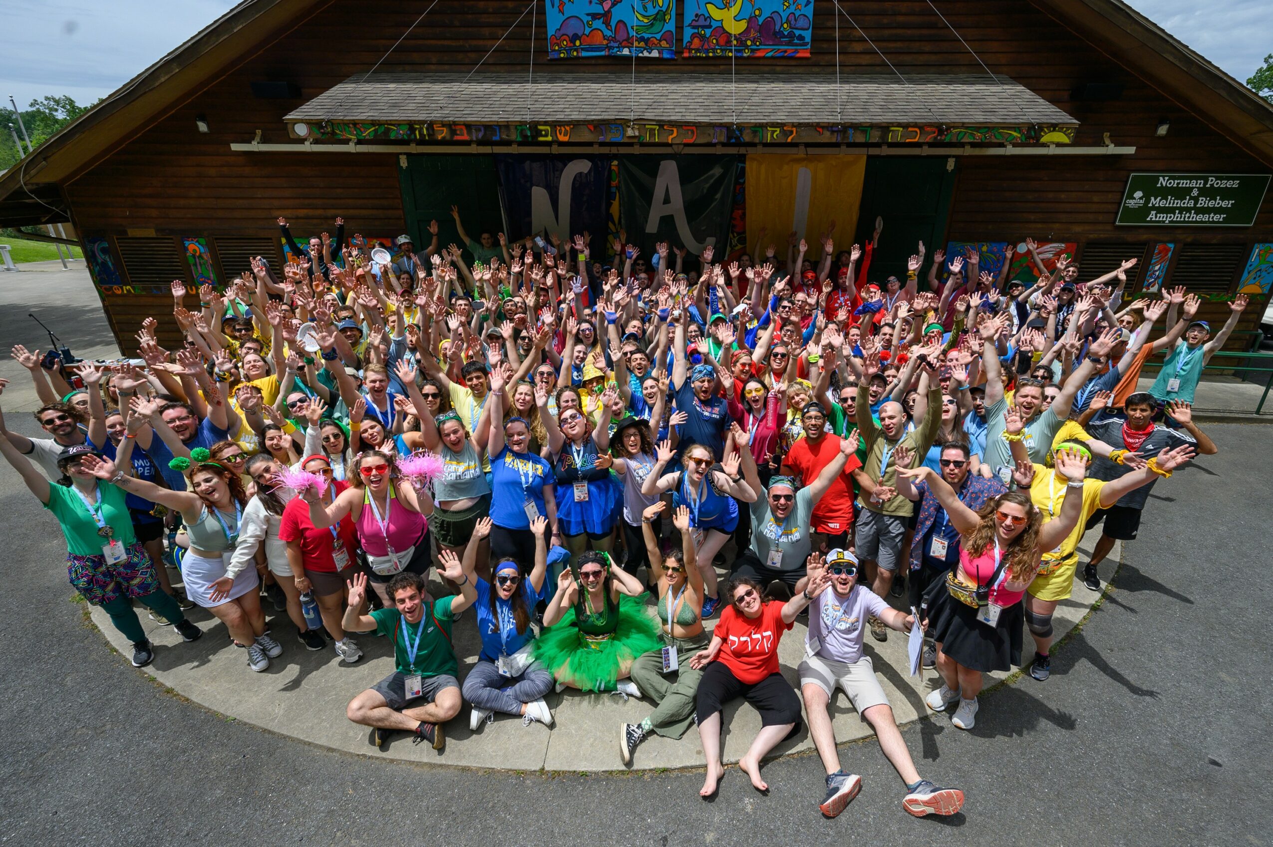 Large group of people gathered outside a building, raising their hands, with a colorful sign above the entrance.