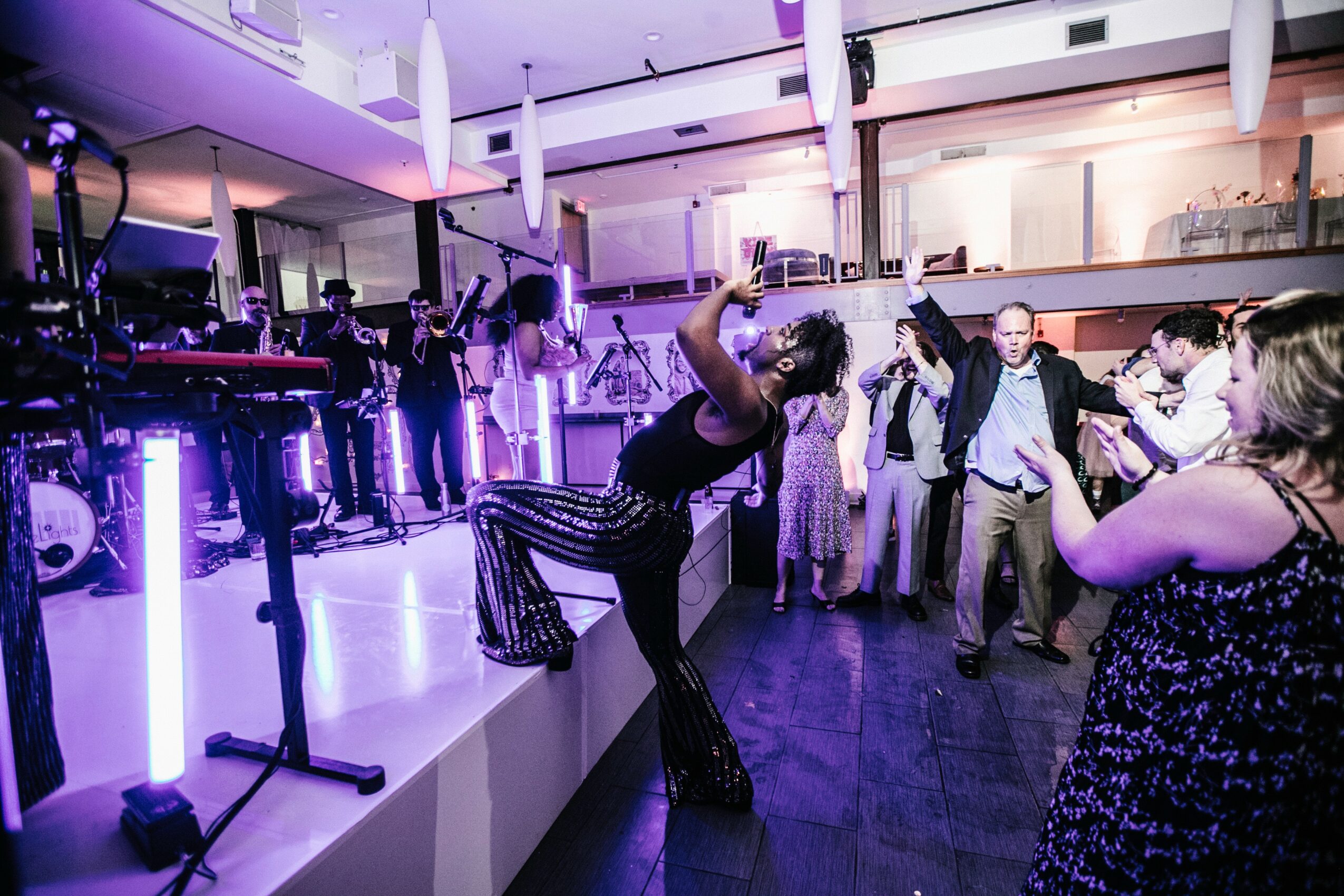 People dancing and socializing in a modern indoor venue with purple lighting and a DJ booth, some taking photos.