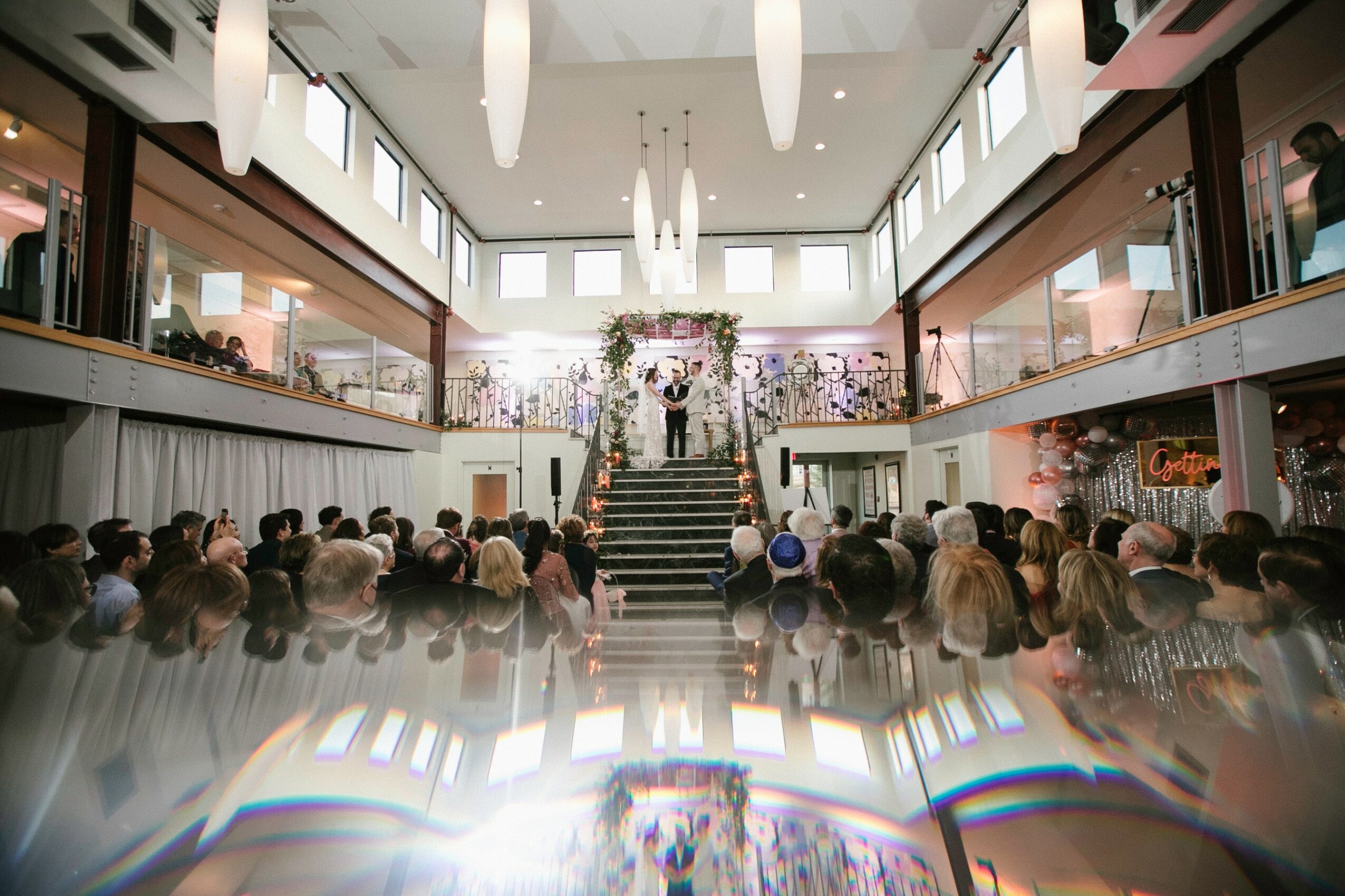 People gathered in a spacious, modern indoor venue with high ceilings, large windows, and a floral arrangement at the front.