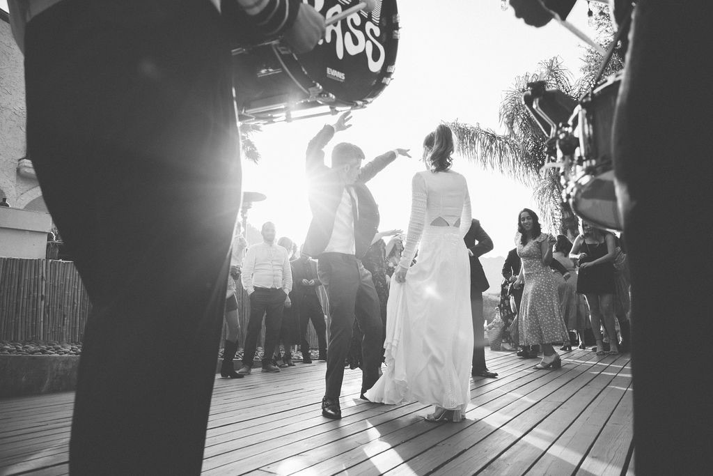 People dancing outdoors with bright light in background, some in formal attire, on a wooden floor, surrounded by onlookers.