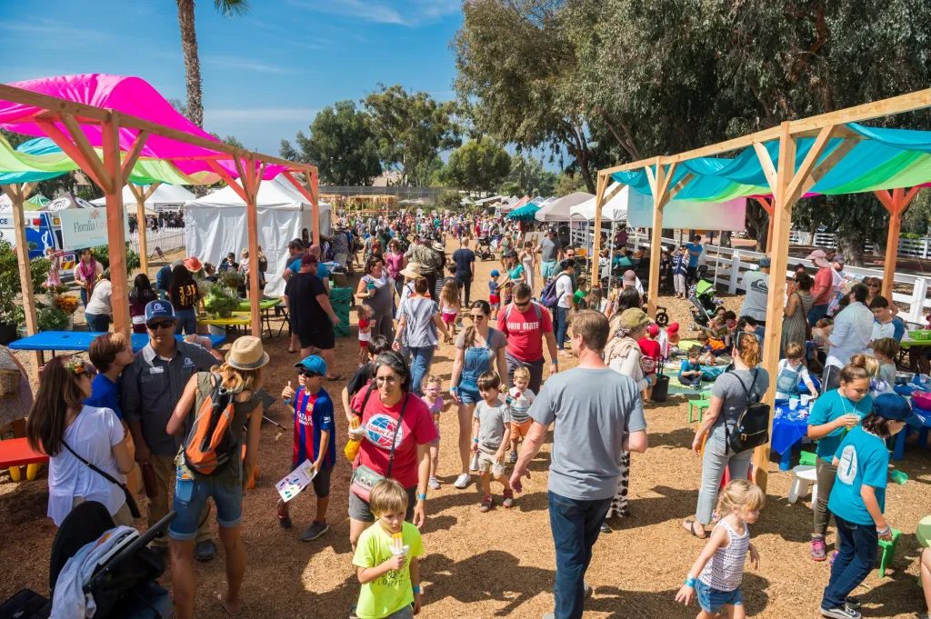 Crowd of people at an outdoor market with colorful tents and trees in the background.