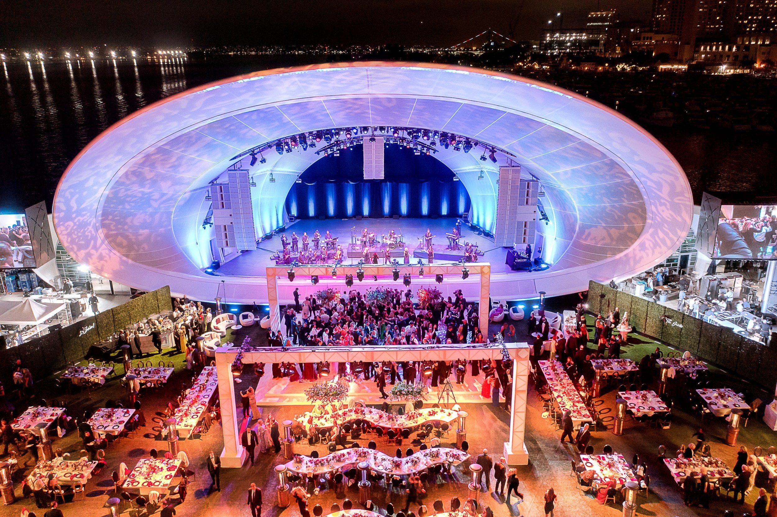 Outdoor concert stage with a large, illuminated, oval-shaped structure, surrounded by tables and people at night.