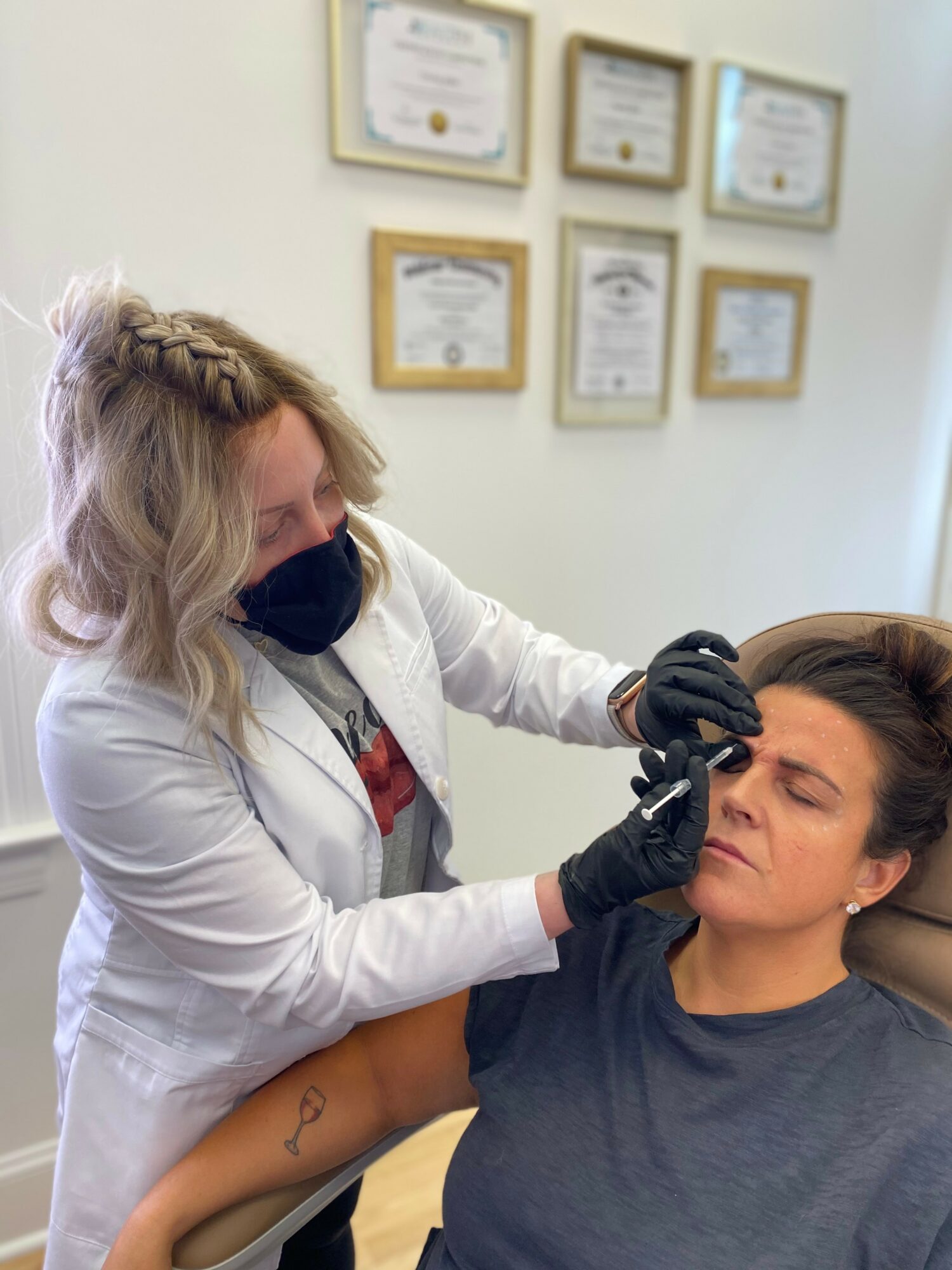 A woman in a white coat and black mask applies a cosmetic treatment to a seated woman's face in a clinic, certificates on the wall.