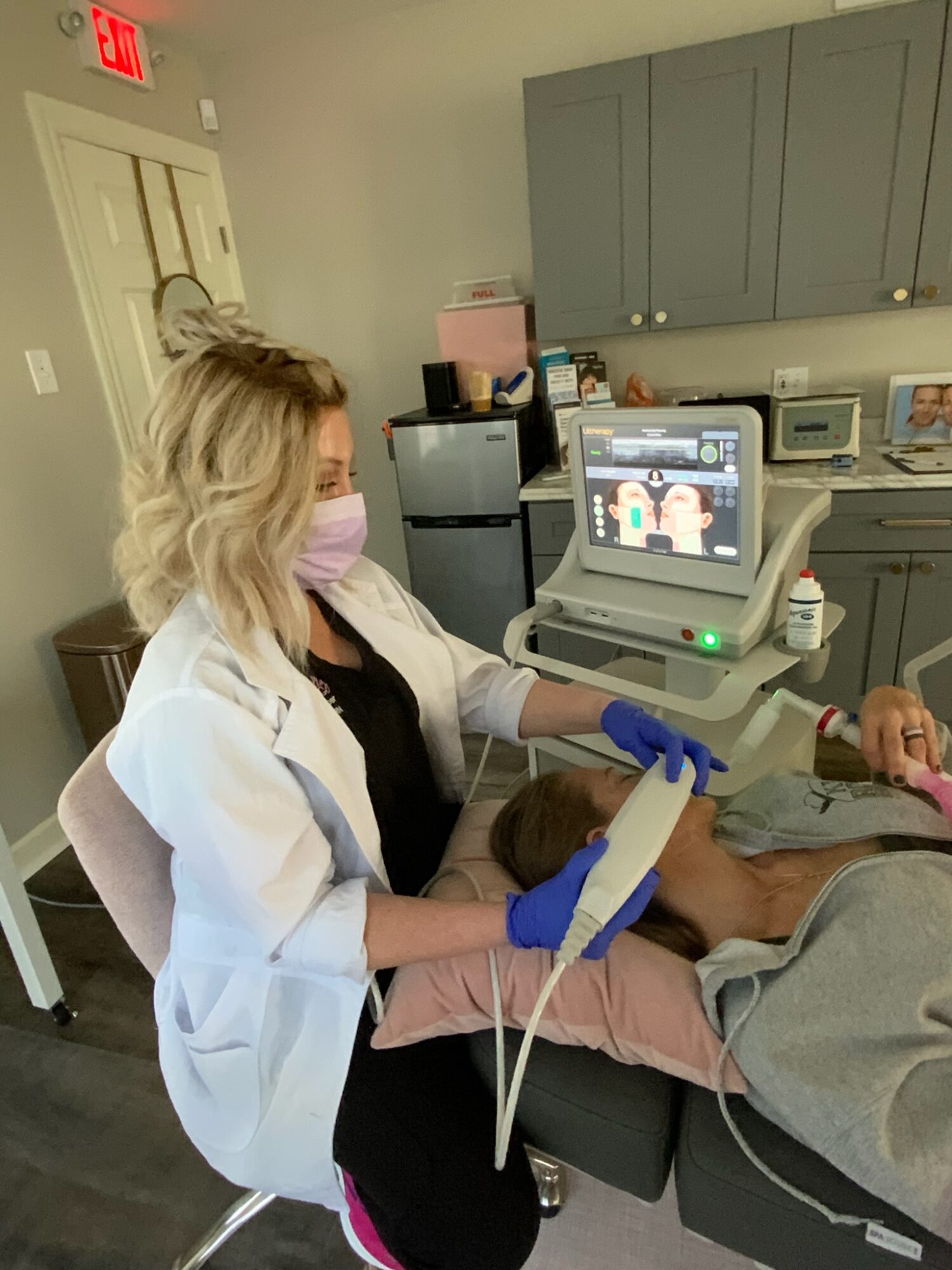 Medical professional performs ultrasound on patient lying on examination table, with ultrasound machine and medical cabinets in background.