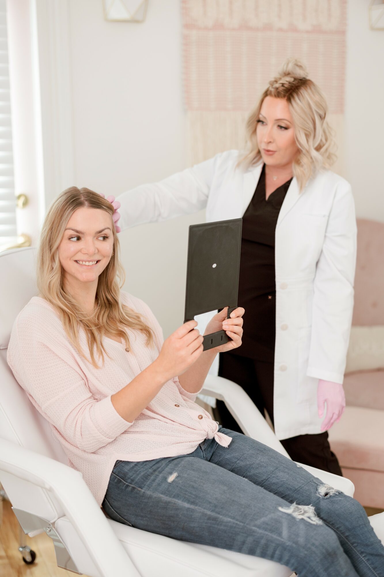 Two women in a medical setting, one sitting and holding a tablet, the other standing with a clipboard, smiling and engaging.