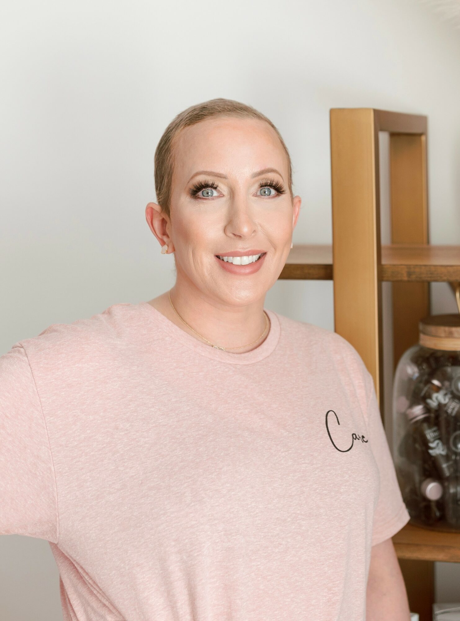 Smiling woman with blonde hair in a pink shirt standing indoors near a wooden shelf with a glass jar.