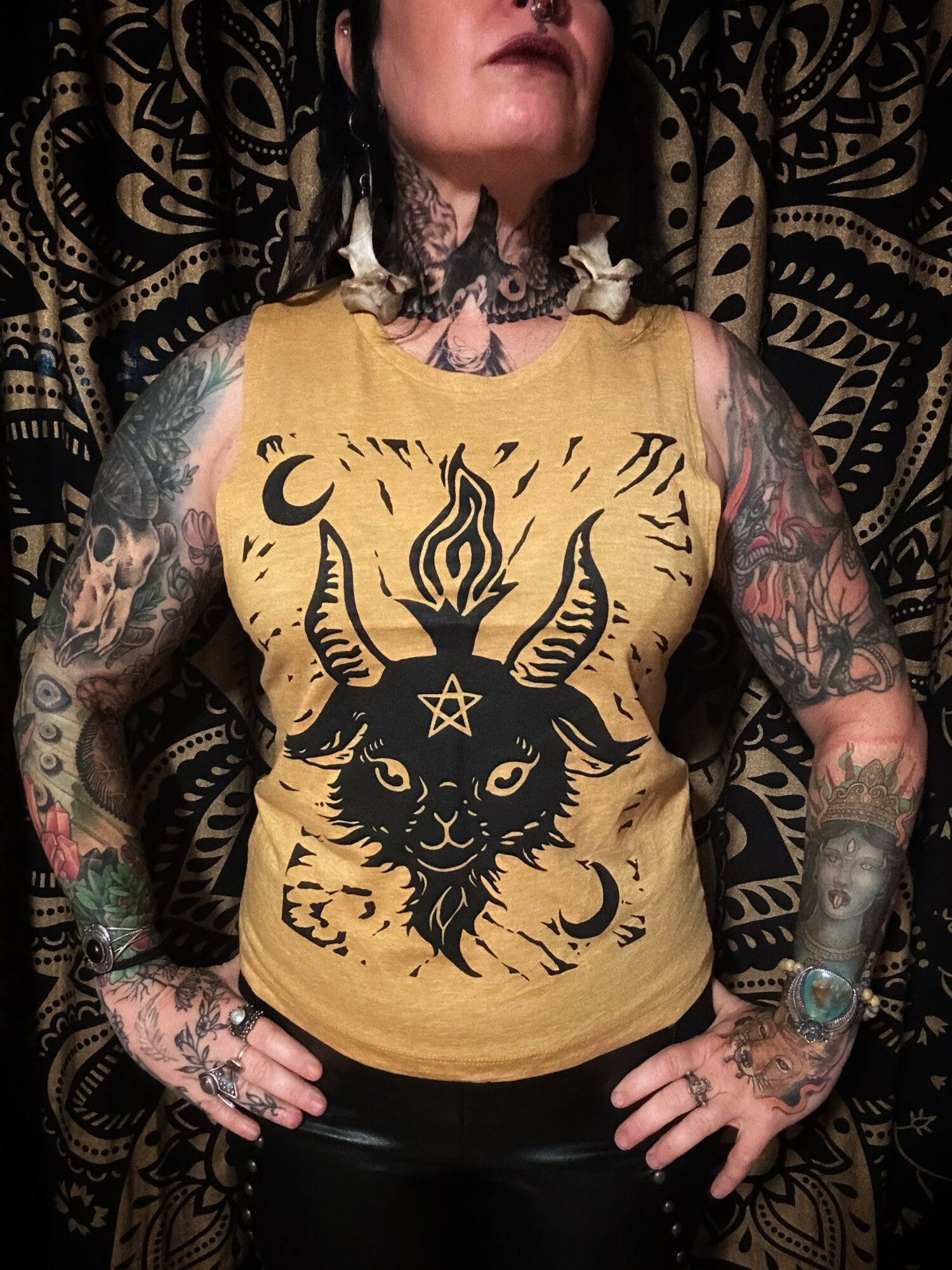 Person with tattoos wearing a yellow tank top with a black goat and pentagram design, standing against a patterned background.