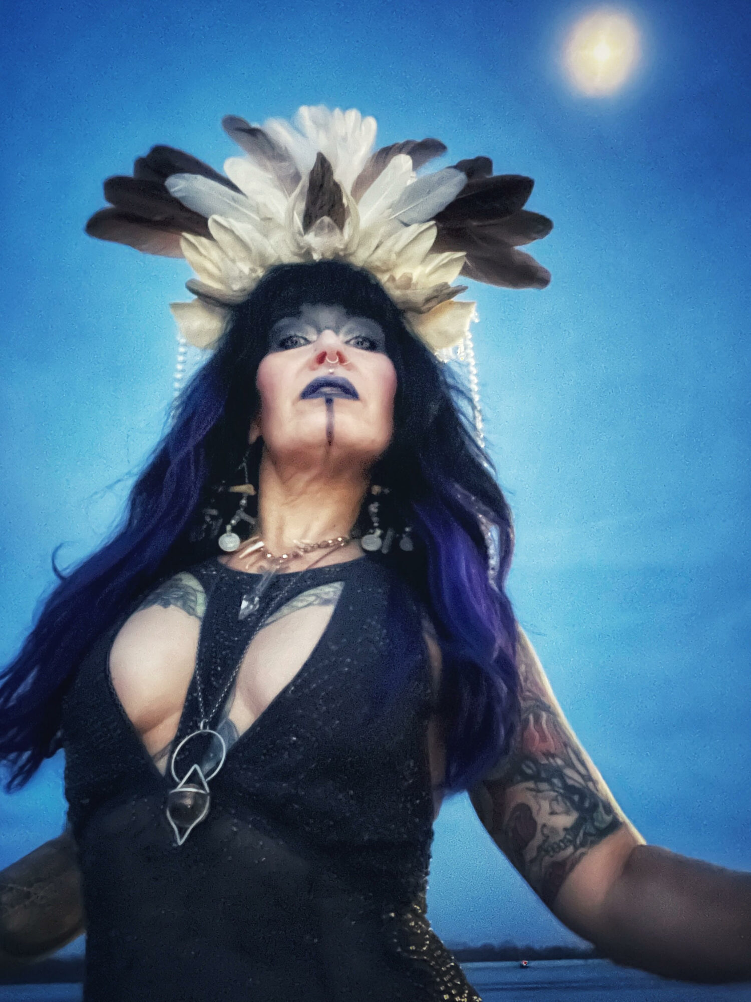 Woman with dark hair and elaborate feather headdress, wearing black dress, standing outdoors at night with moon in sky.