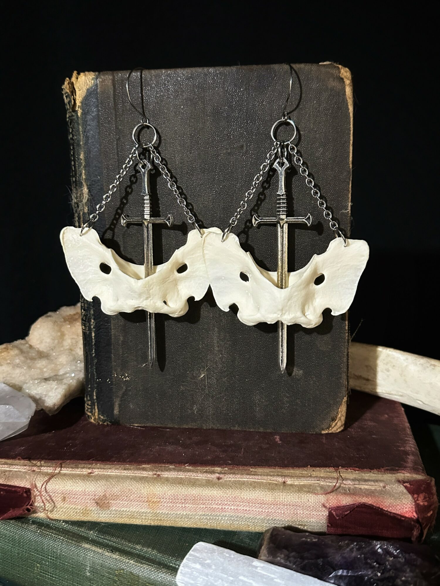 Pair of earrings with cross and butterfly-shaped elements hanging from chains, displayed on a black book with other books and objects around.