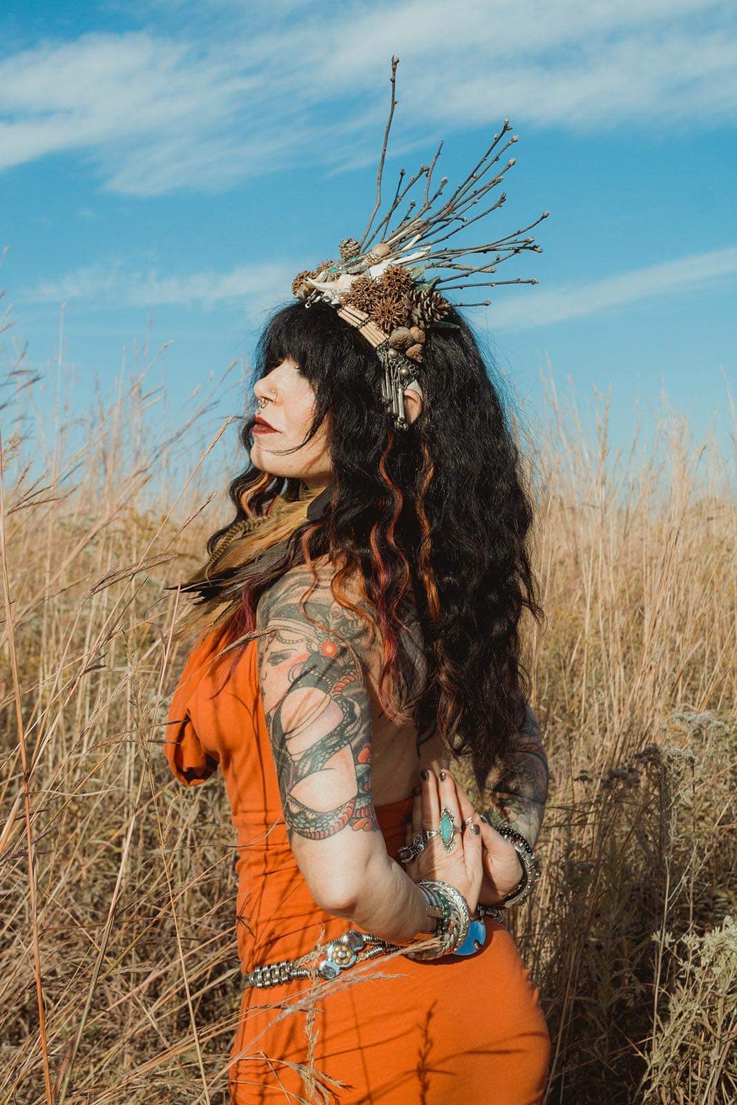 Woman with long curly hair wearing an orange dress and a decorative headpiece stands in a field of tall grass under a blue sky.