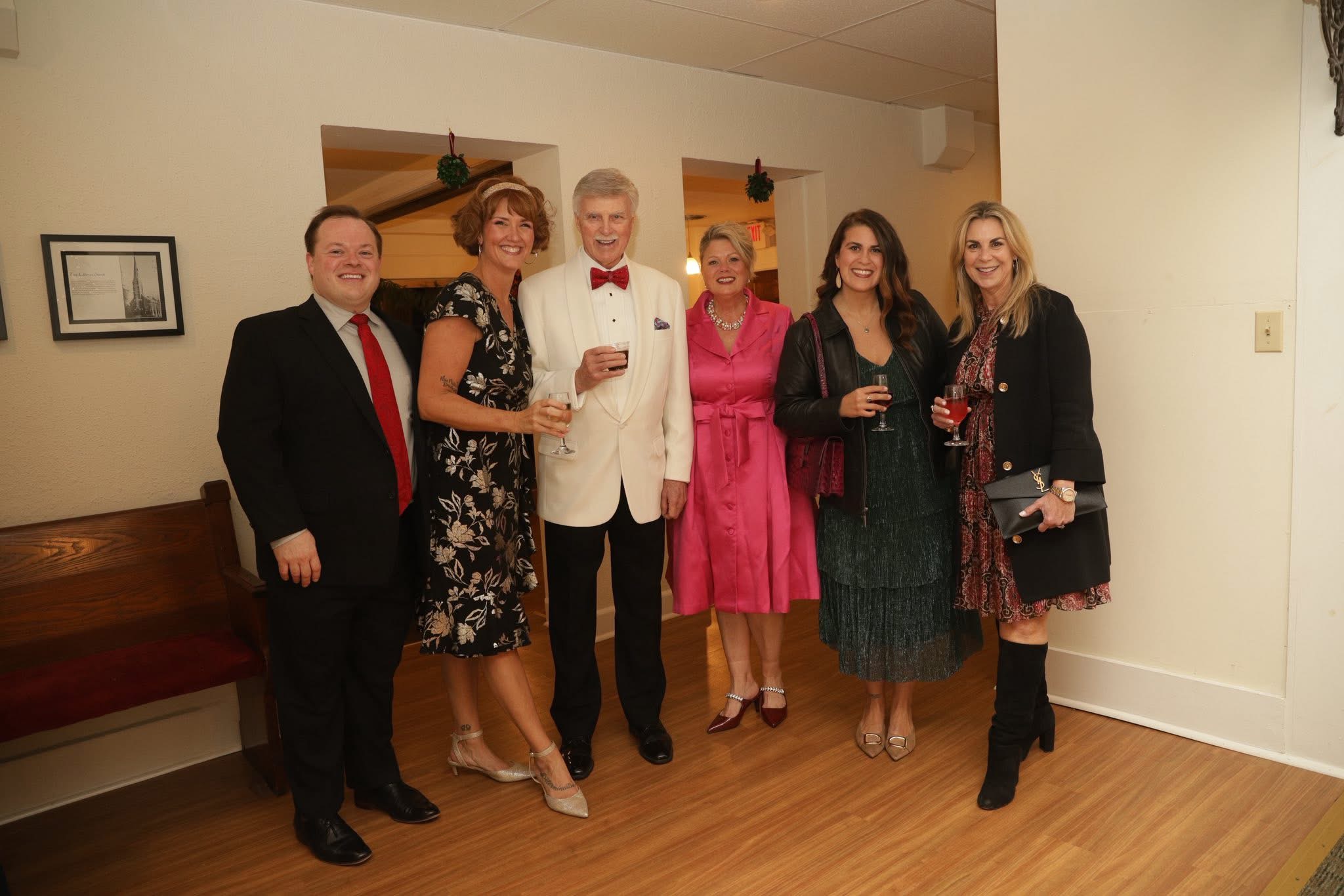 Group of seven people dressed in formal attire, standing indoors, smiling, holding drinks, with a wooden floor and white walls.