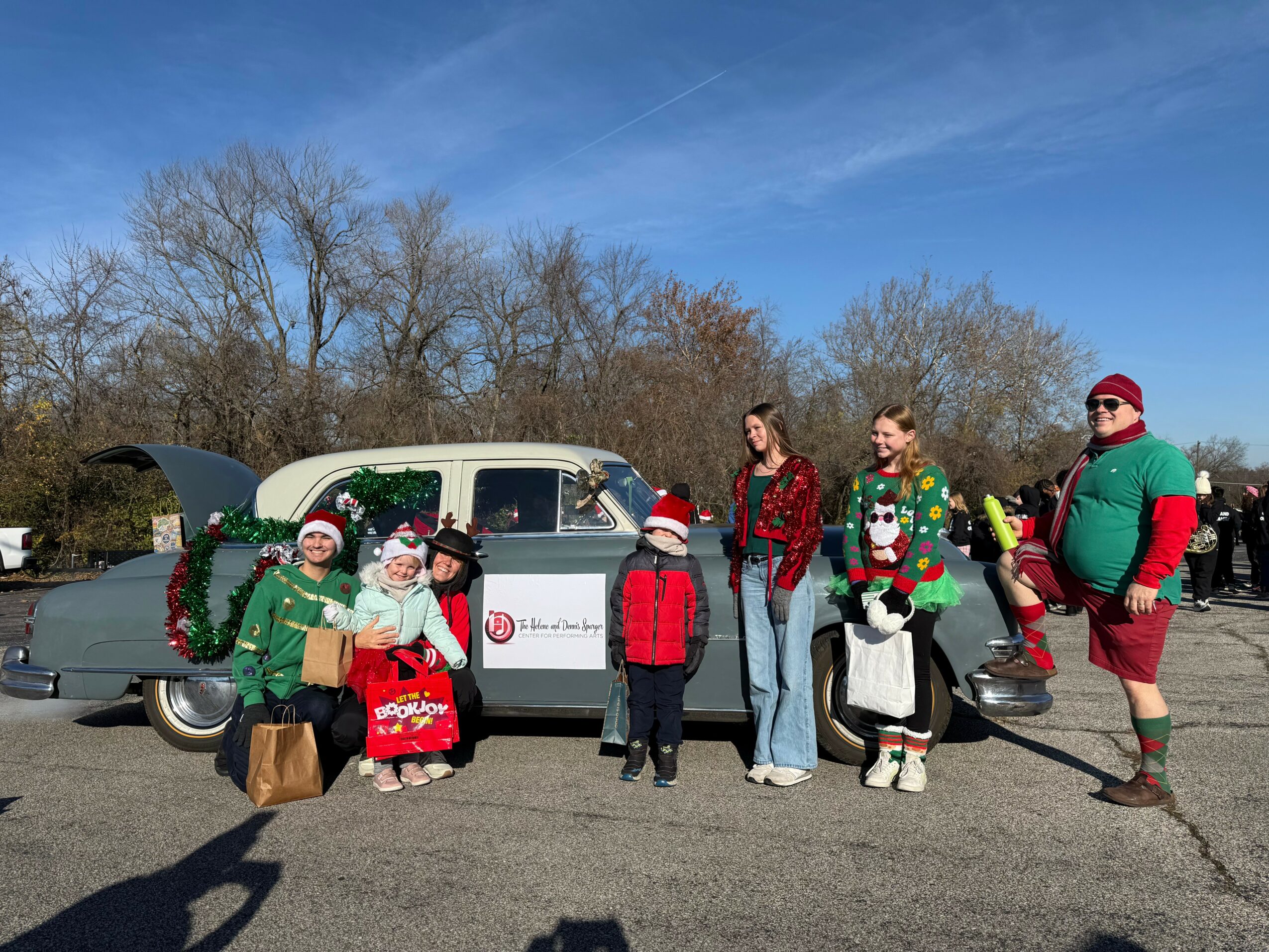 Group of people in Christmas attire standing next to a vintage car outdoors, with trees and a blue sky in background.