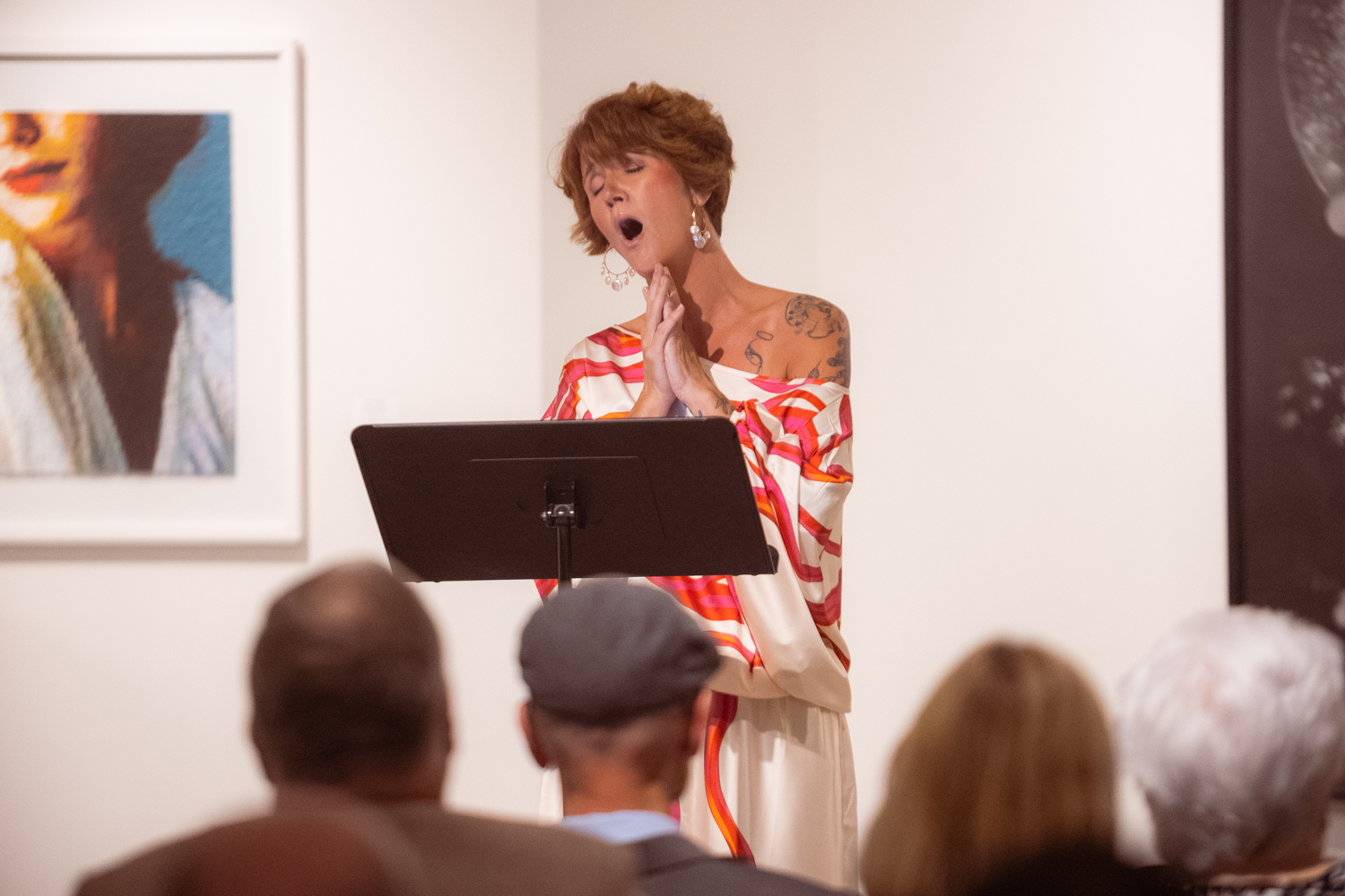 A woman with short hair and tattoos sings passionately with eyes closed, standing in front of a music stand, in an art gallery.