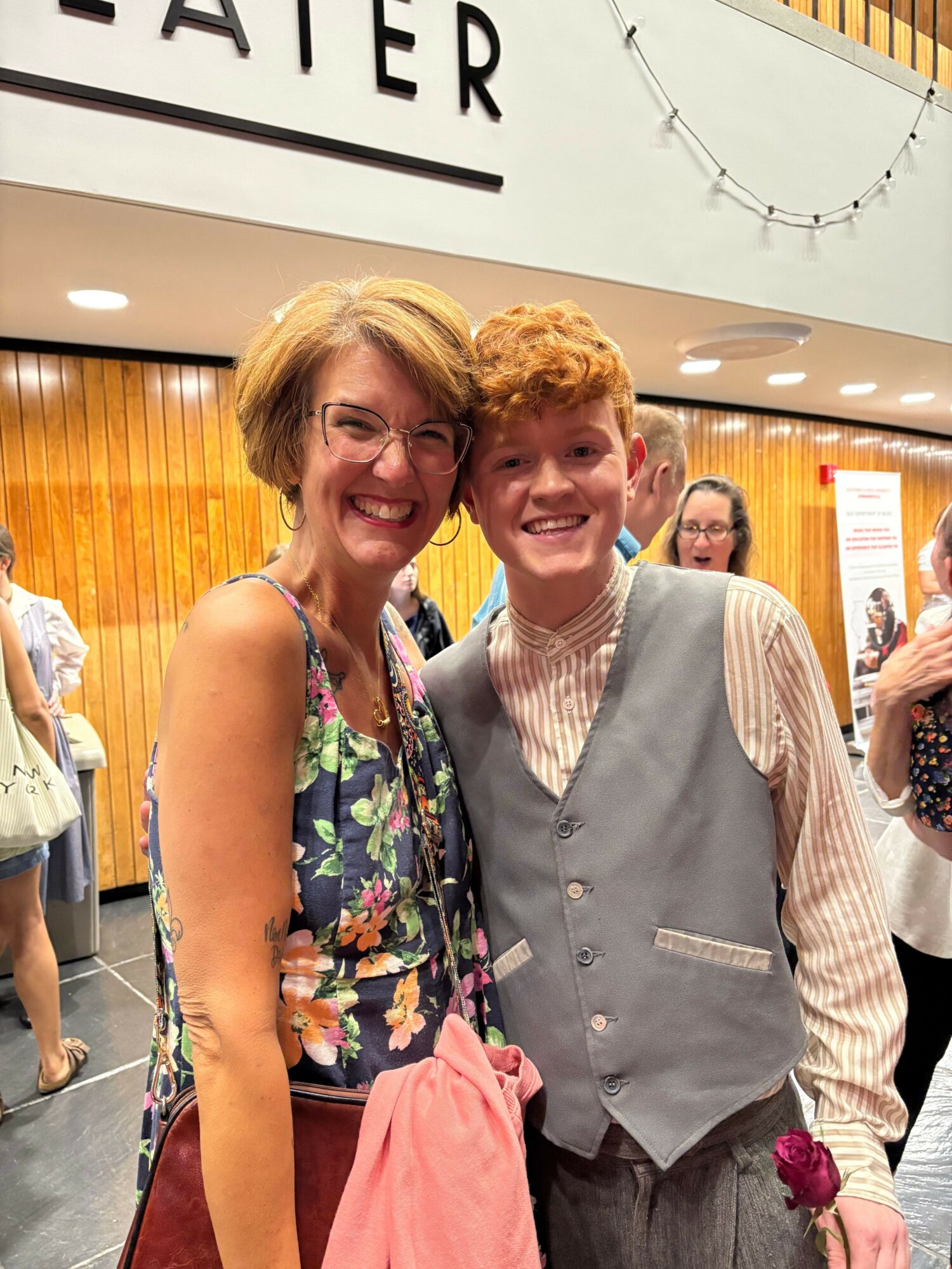 Two women smiling and posing for a photo indoors, with a wooden wall and a white sign in the background.