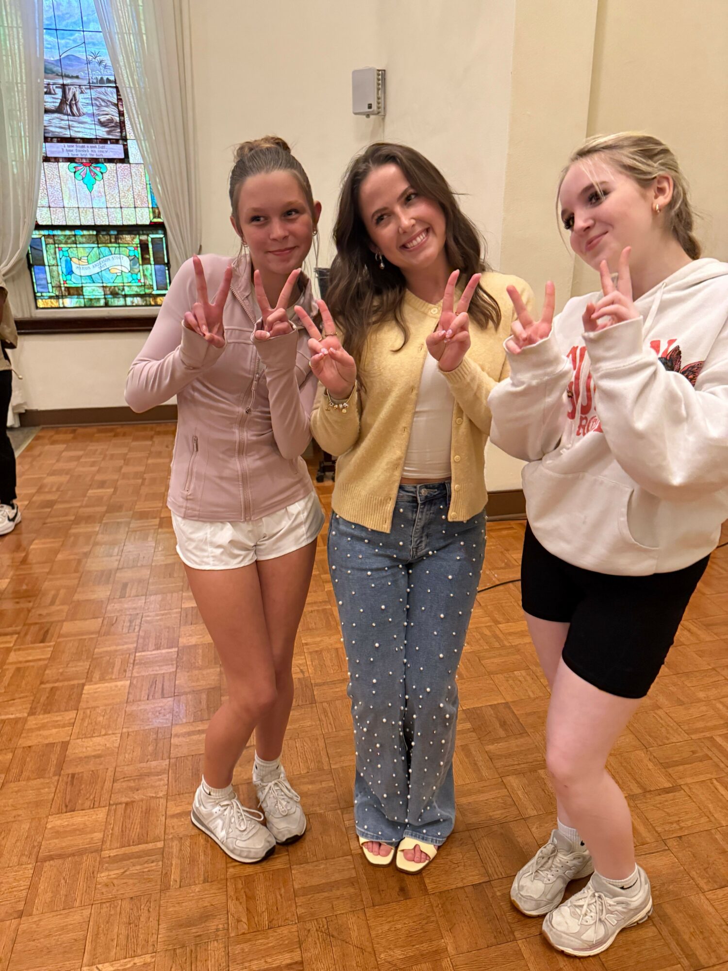 Three young women posing indoors, making peace signs, standing on a wooden floor, with a stained glass window in the background.